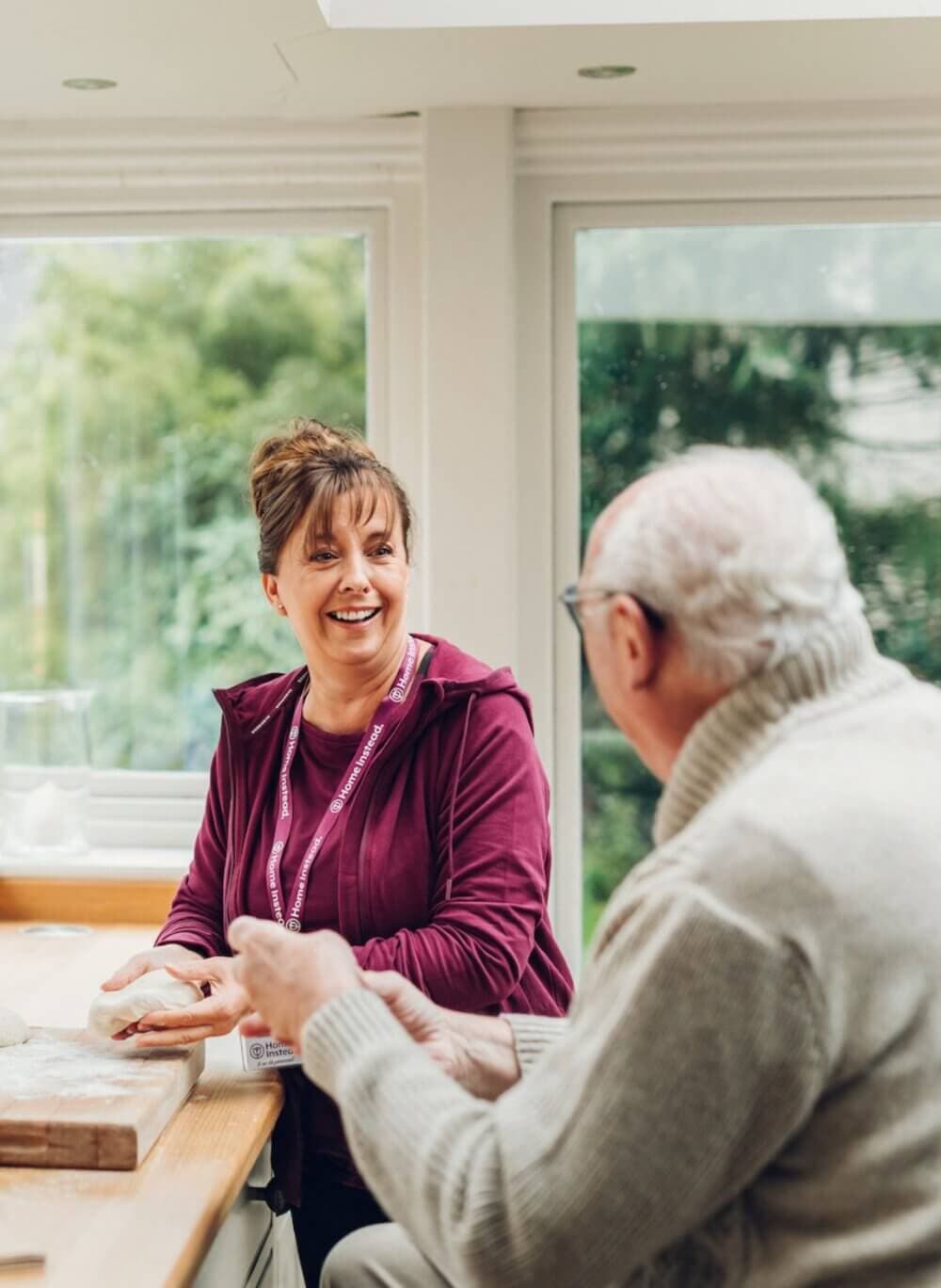 A caregiver smiles and talks with an elderly man at a kitchen table, with large windows providing a view of greenery. - Home Instead