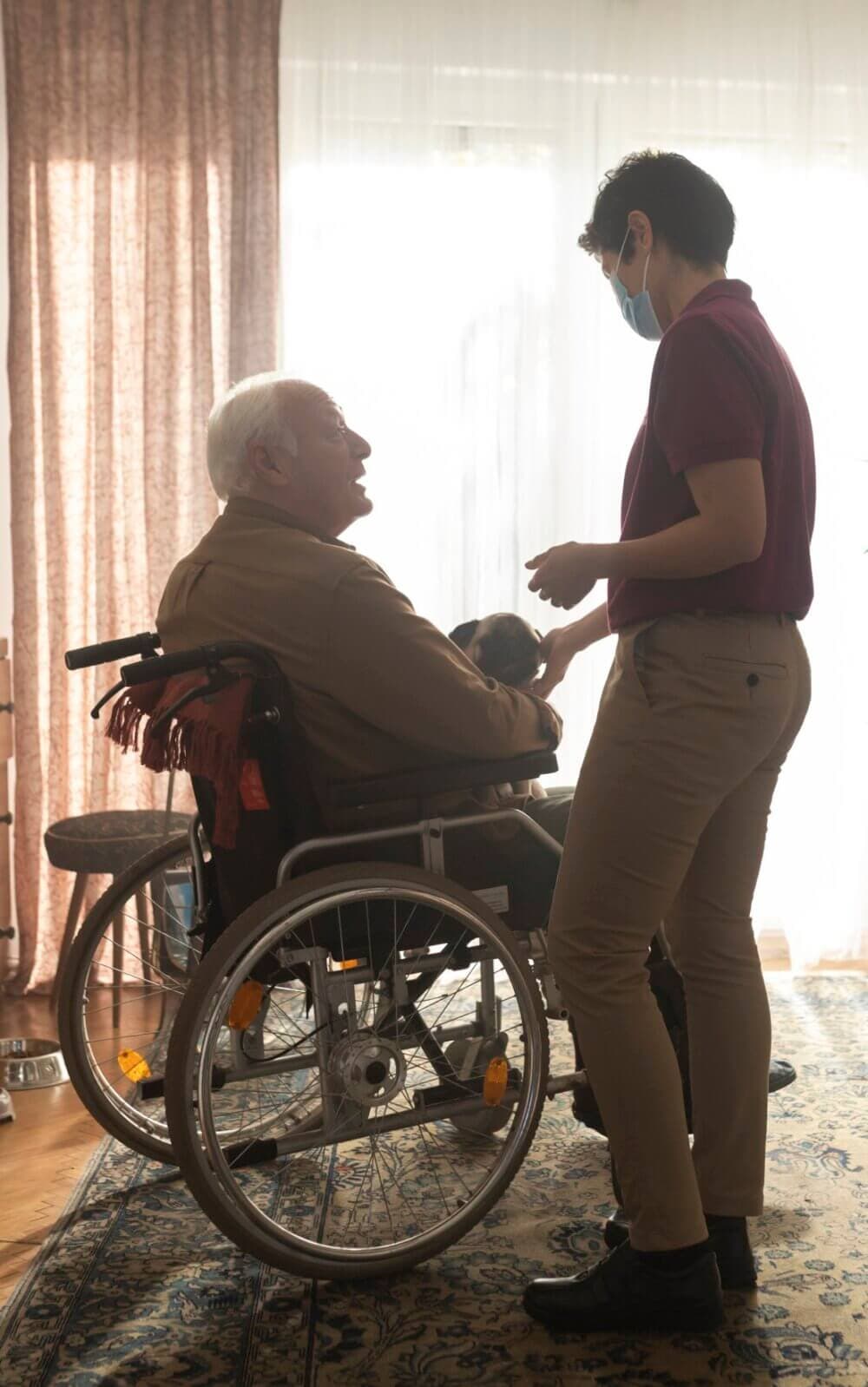 A caregiver assists an elderly man in a wheelchair holding a small dog, both by a sunlit window. - Home Instead