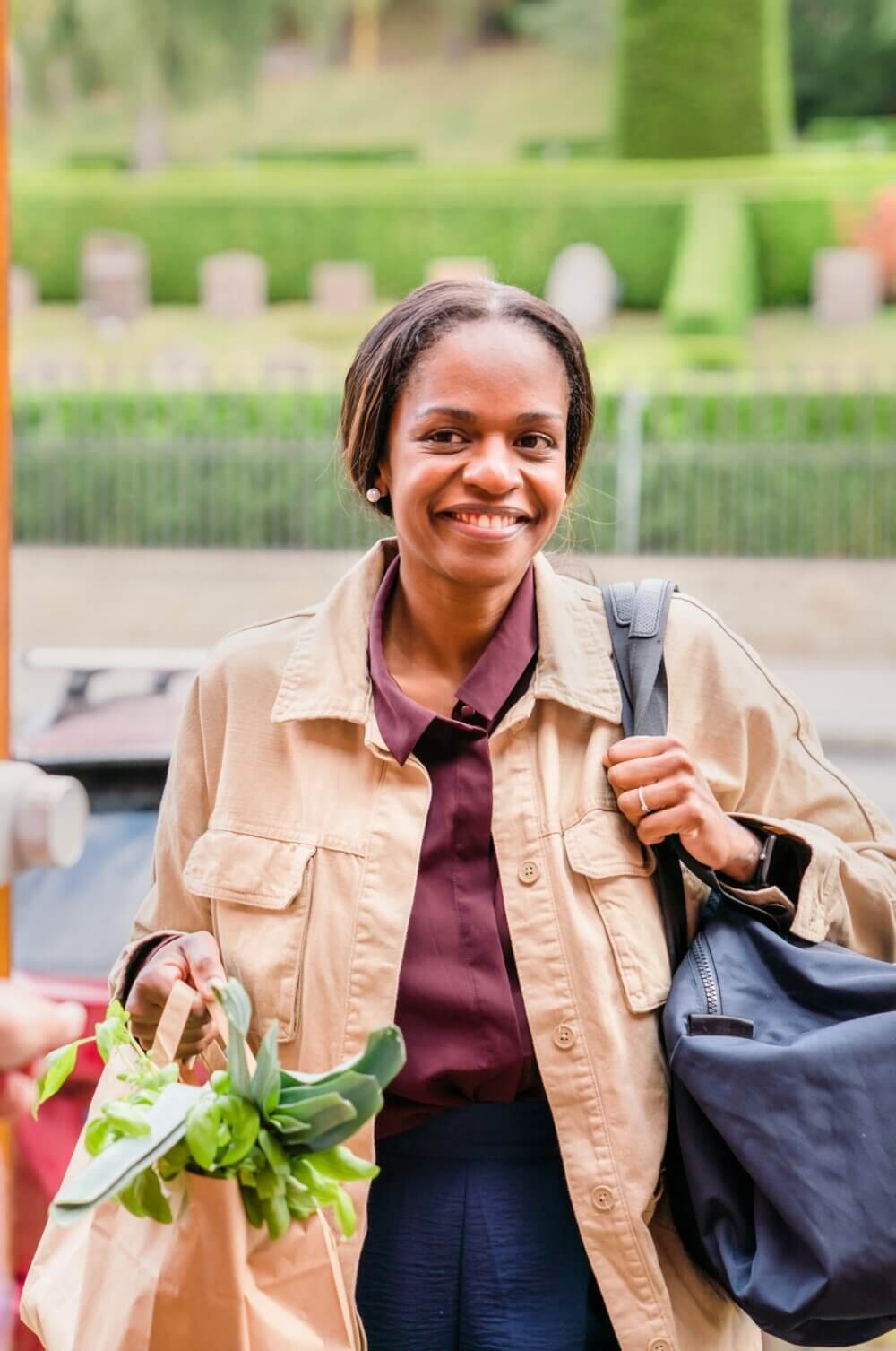 A smiling person with a shopping bag and shoulder bag stands outdoors, wearing a beige jacket and maroon shirt. - Home Instead
