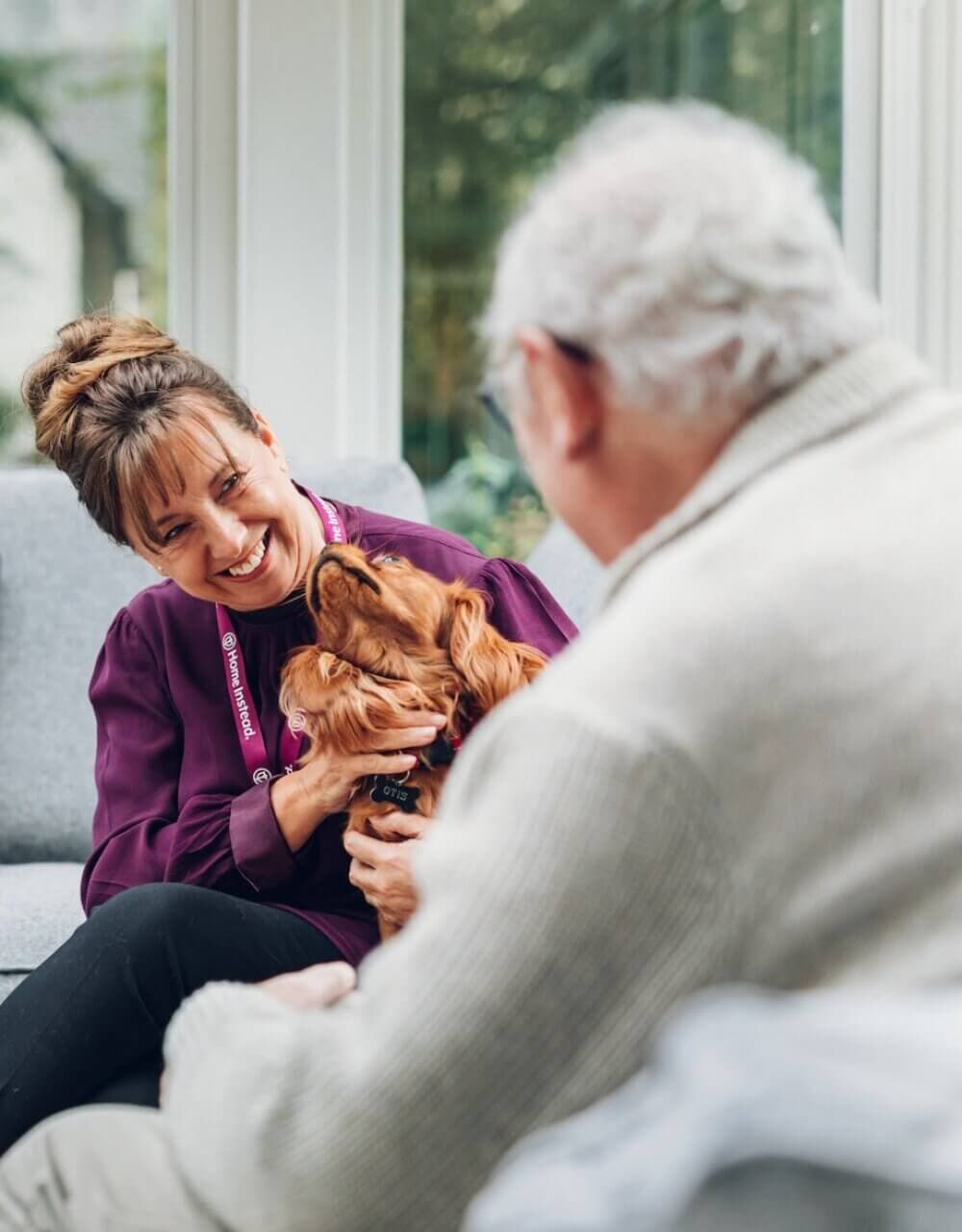 A woman and an elderly man smile while petting a brown dog indoors. - Home Instead