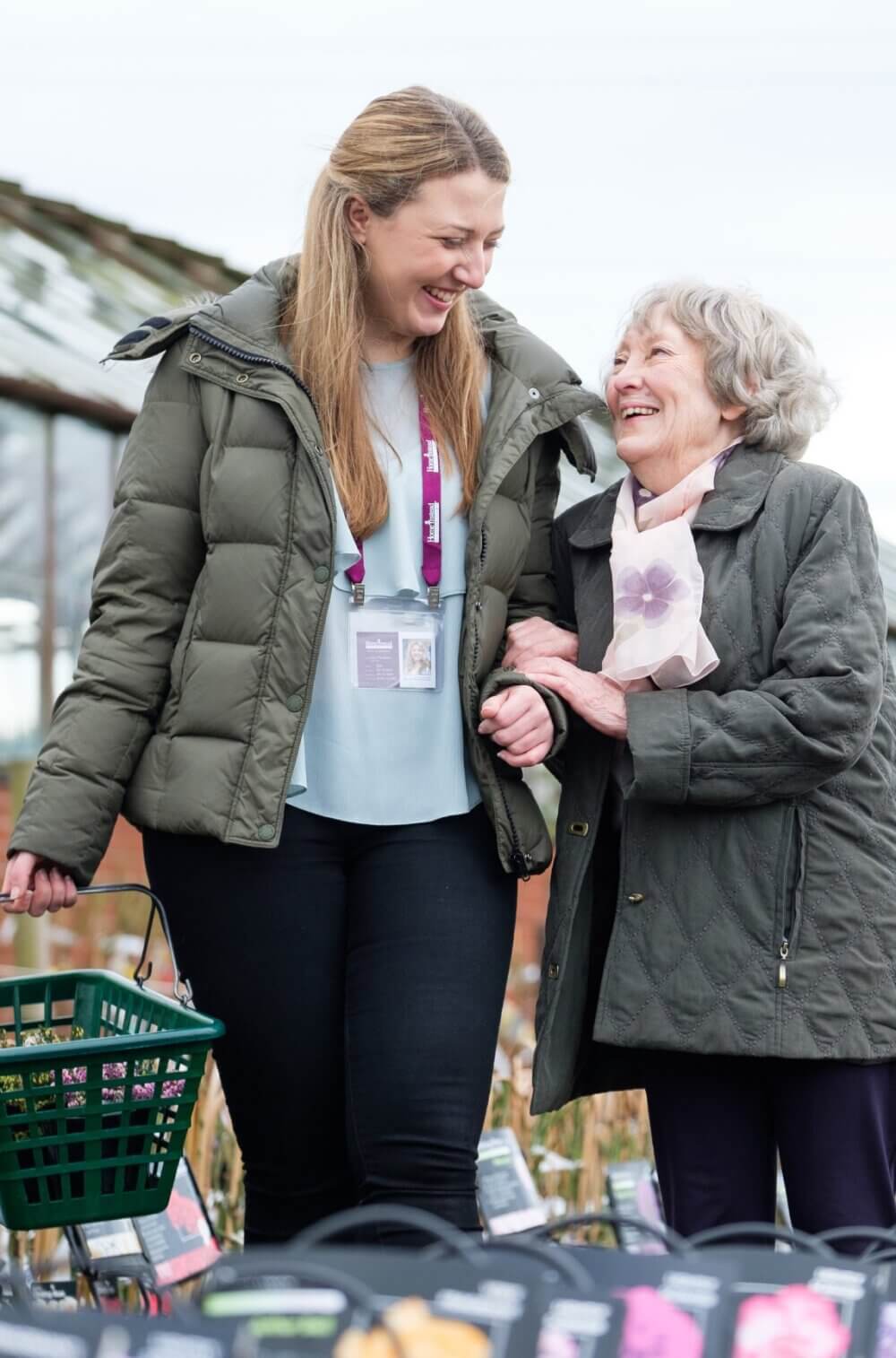 A young woman and an elderly woman smile and walk arm-in-arm outside while holding a shopping basket. - Home Instead