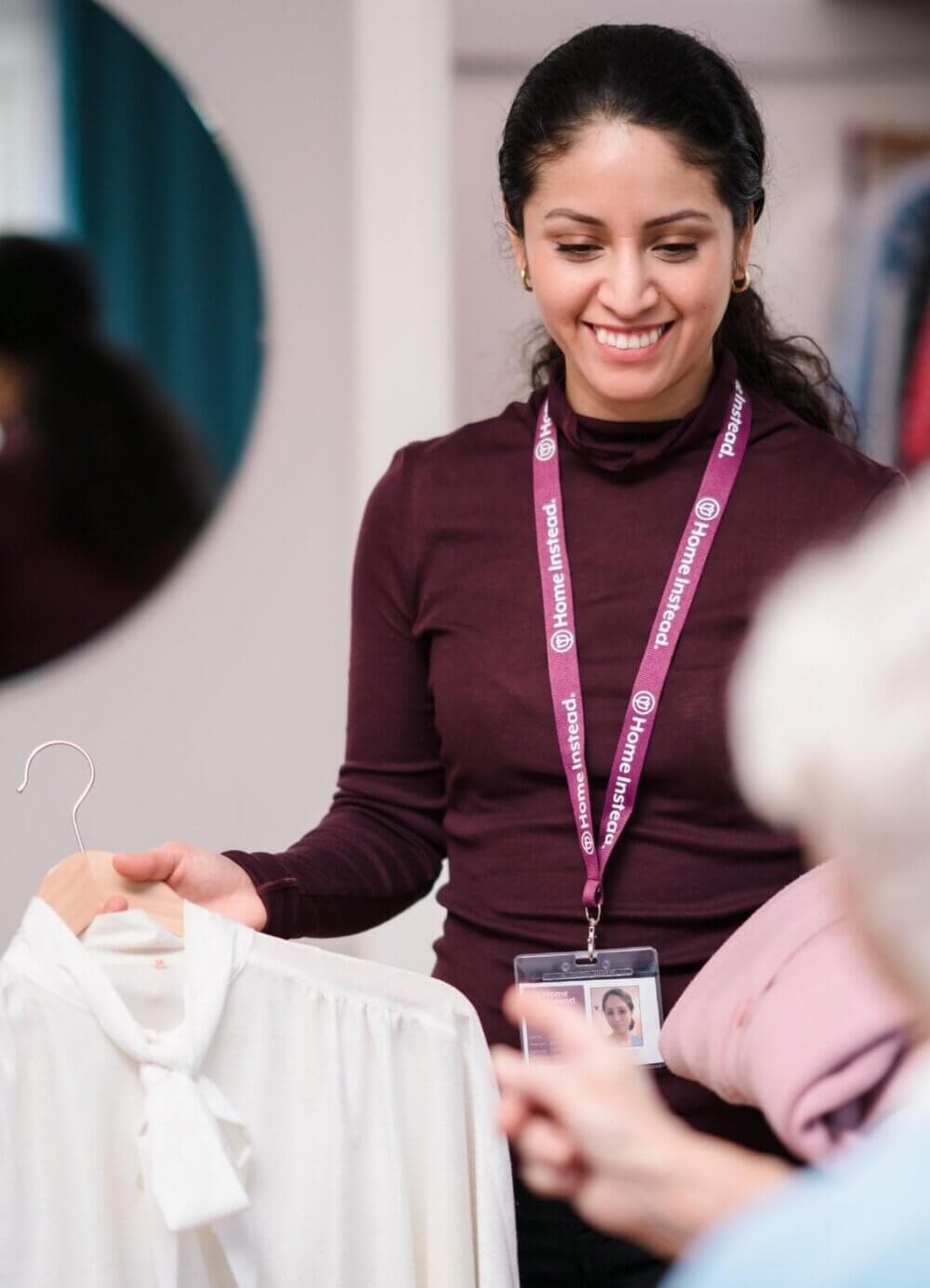 A woman with a lanyard helps a customer by holding up a white blouse on a hanger in a clothing store. - Home Instead