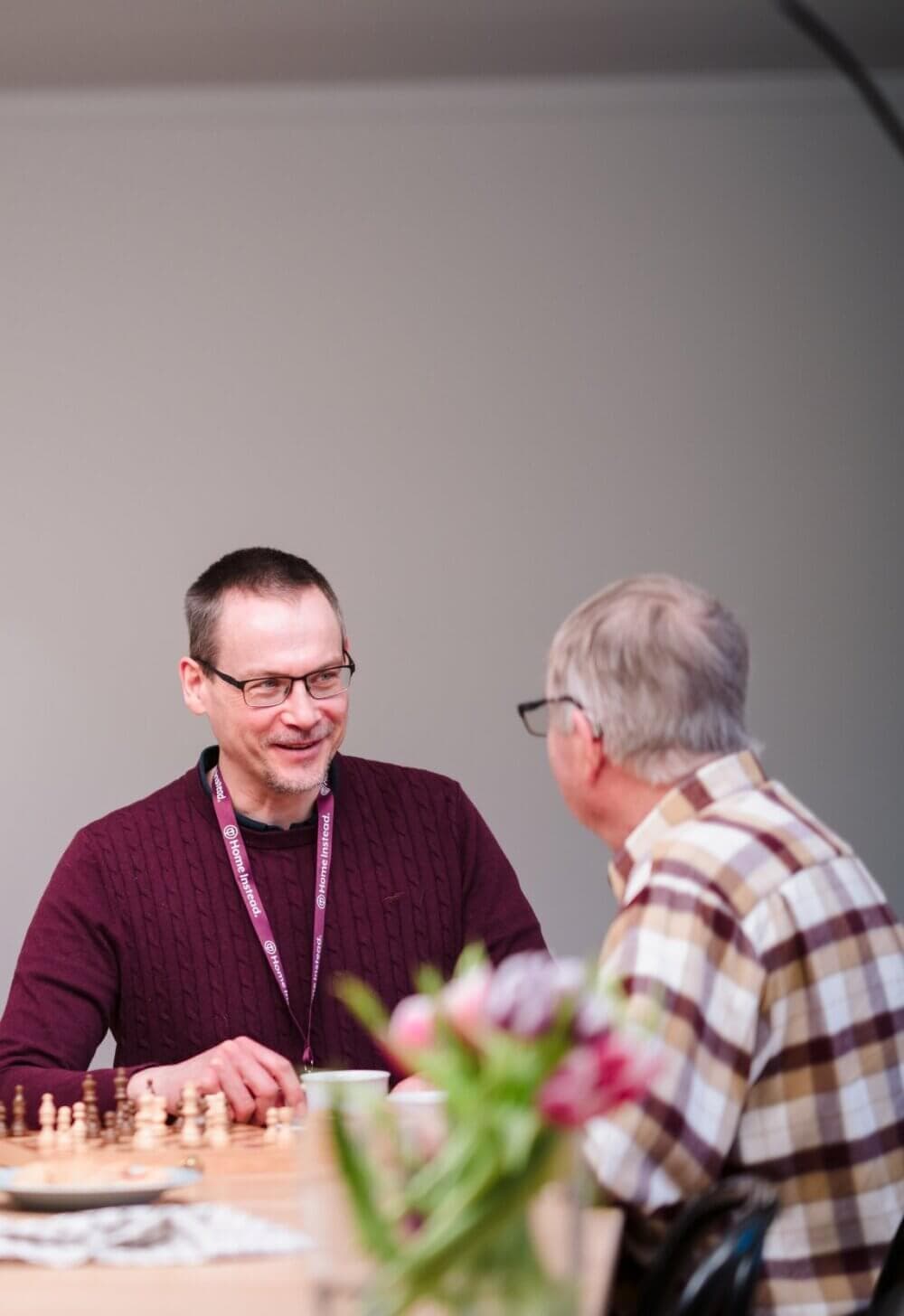Two men conversing at a table with a chessboard and flowers in the foreground. - Home Instead