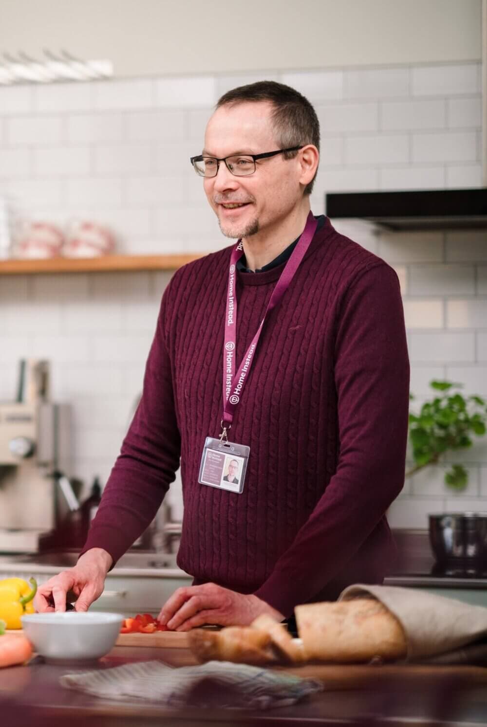 Man wearing glasses and a maroon sweater prepares food in a kitchen, with a lanyard around his neck. - Home Instead