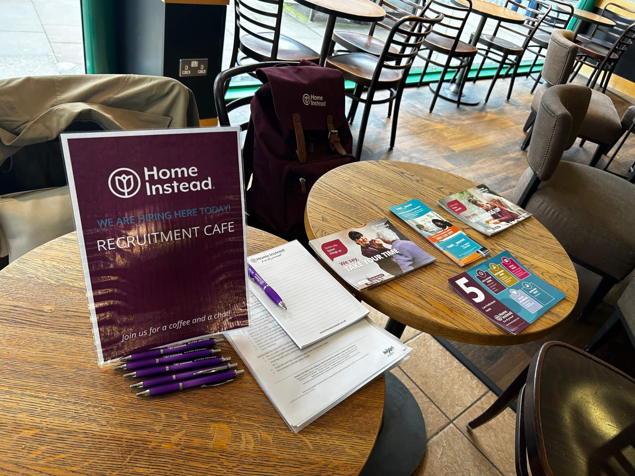 Recruitment event setup at a cafe with brochures, pens, signs, and forms on tables, promoting hiring opportunities. - Home Instead