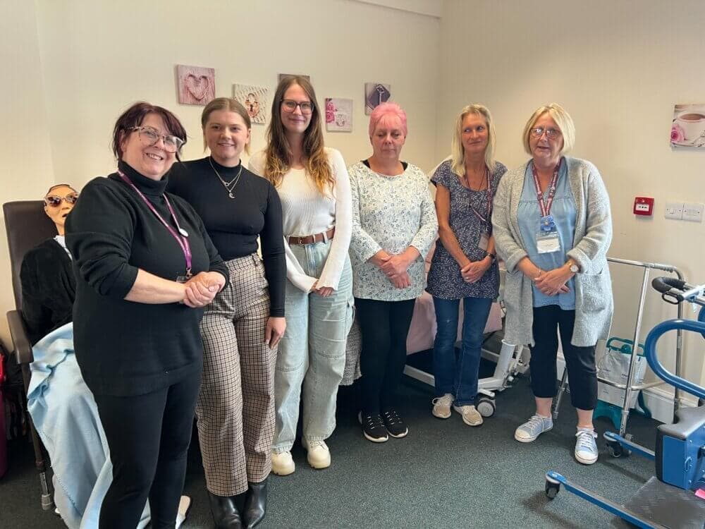 A group of six women standing in a room, smiling at the camera with medical equipment visible in the background. - Home Instead