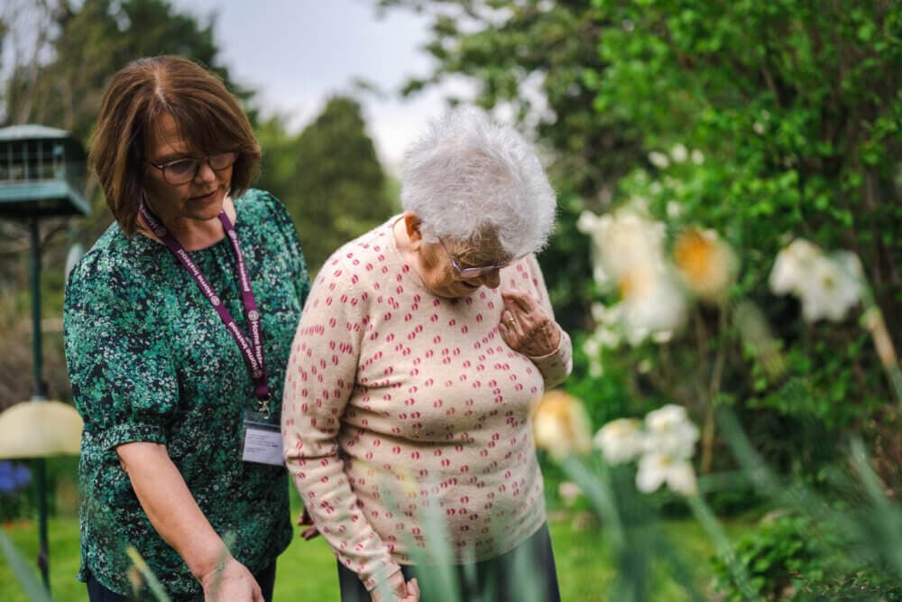 A caregiver assists an elderly woman in a garden, surrounded by greenery and flowers. - Home Instead