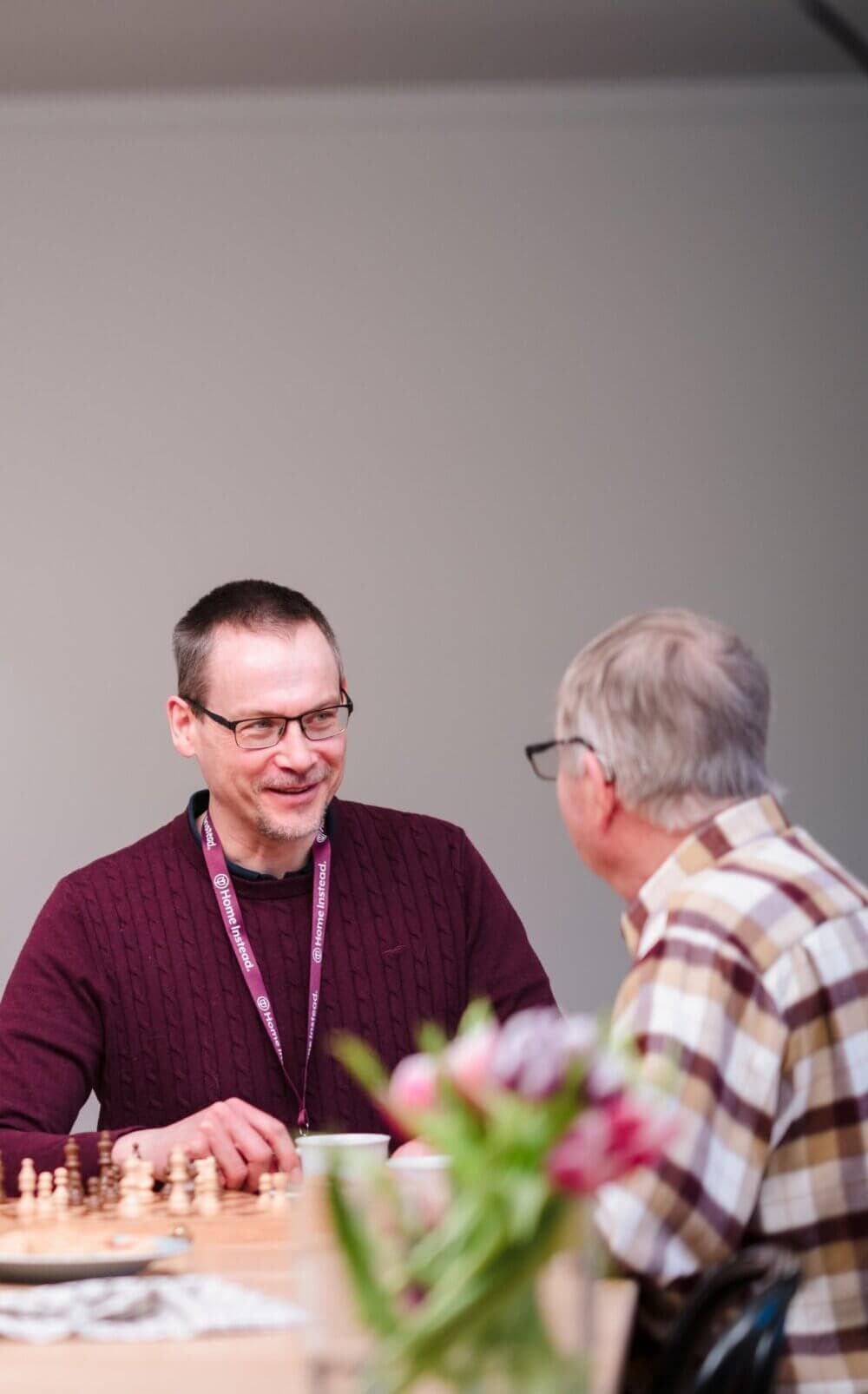 An elderly woman in a maroon jacket smiles while talking with another person, who is holding a watering can. - Home Instead