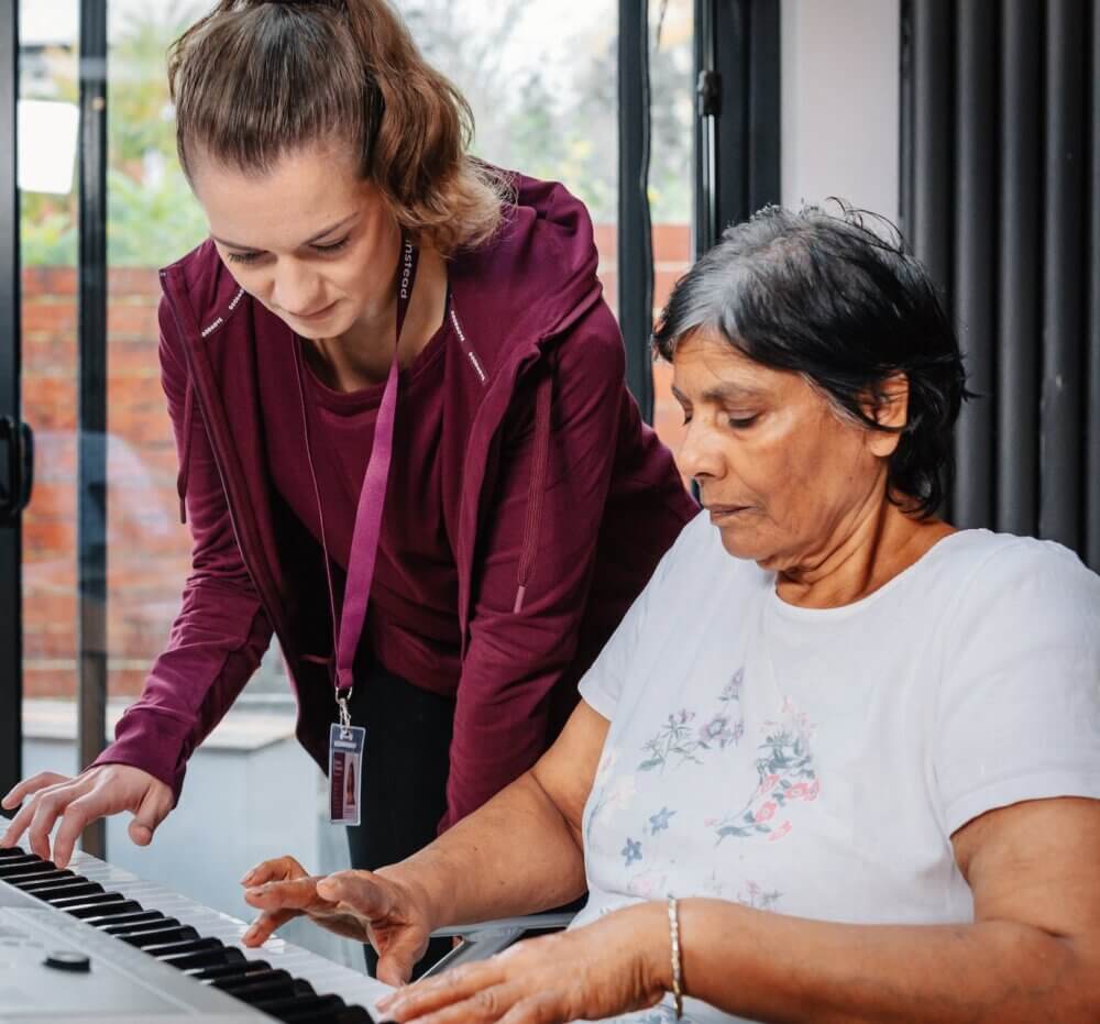A young woman helps an older woman play a keyboard in a brightly lit room. - Home Instead