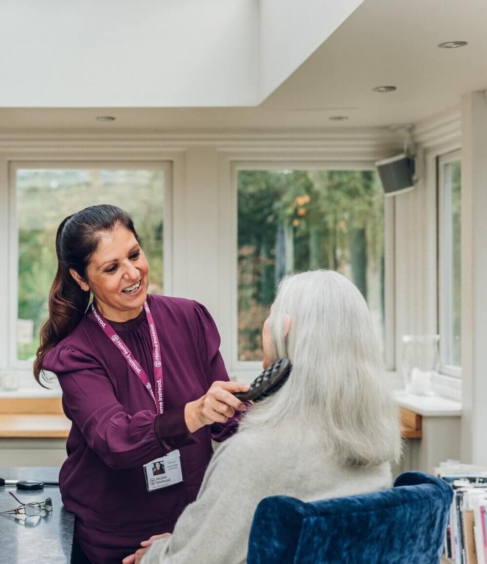 A caregiver in a purple blouse is smiling and brushing the long white hair of an elderly person in a cozy room. - Home Instead