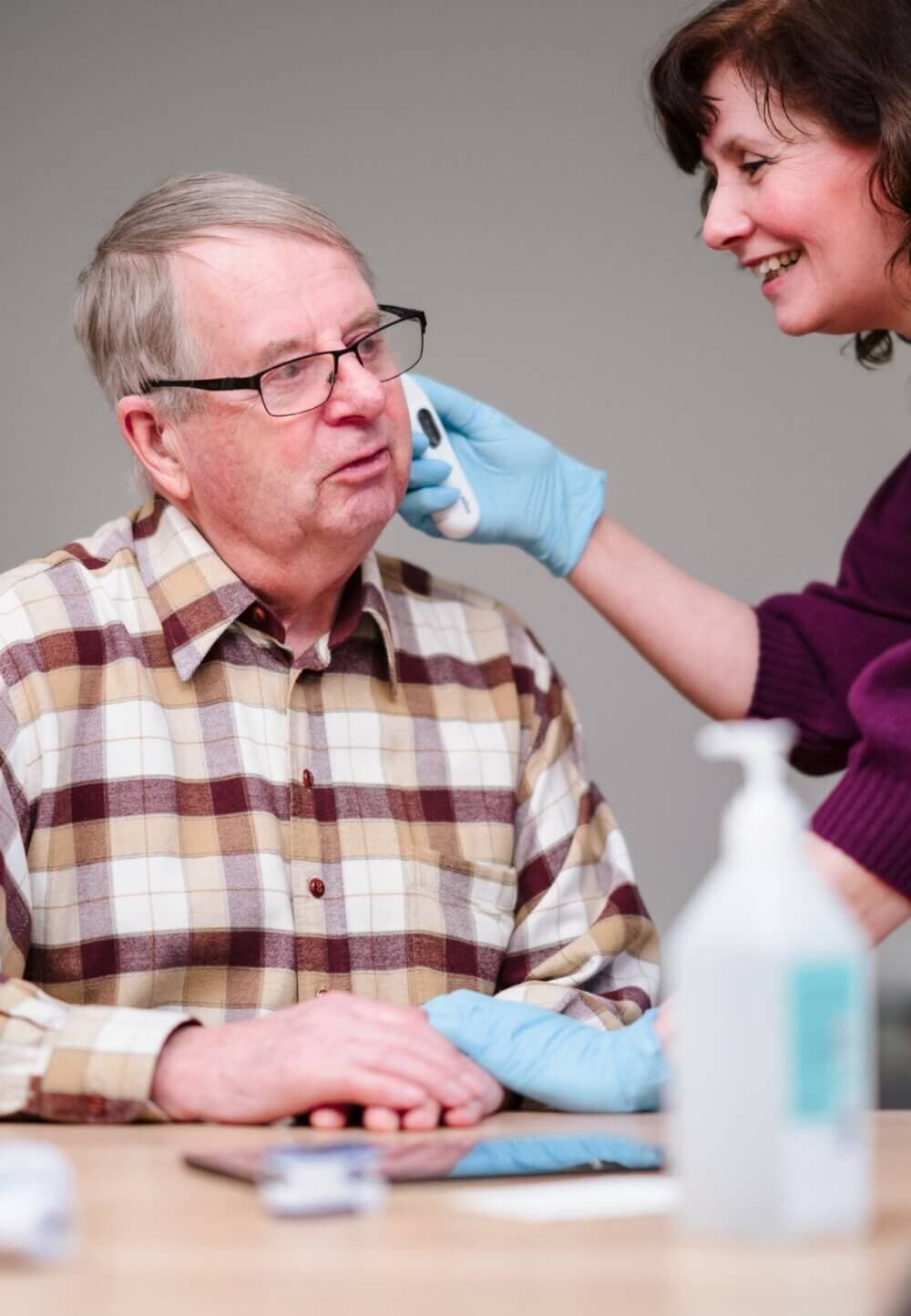 A healthcare worker checks an older man's temperature with an ear thermometer. - Home Instead