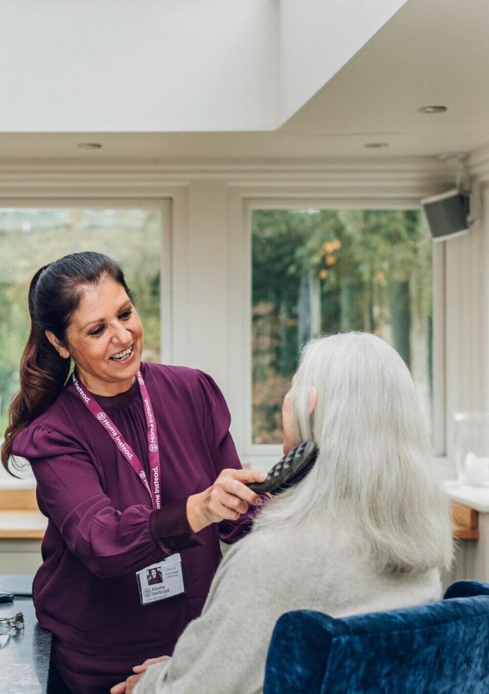 A caregiver smiles while gently brushing an elderly woman's hair in a bright, sunlit room. - Home Instead