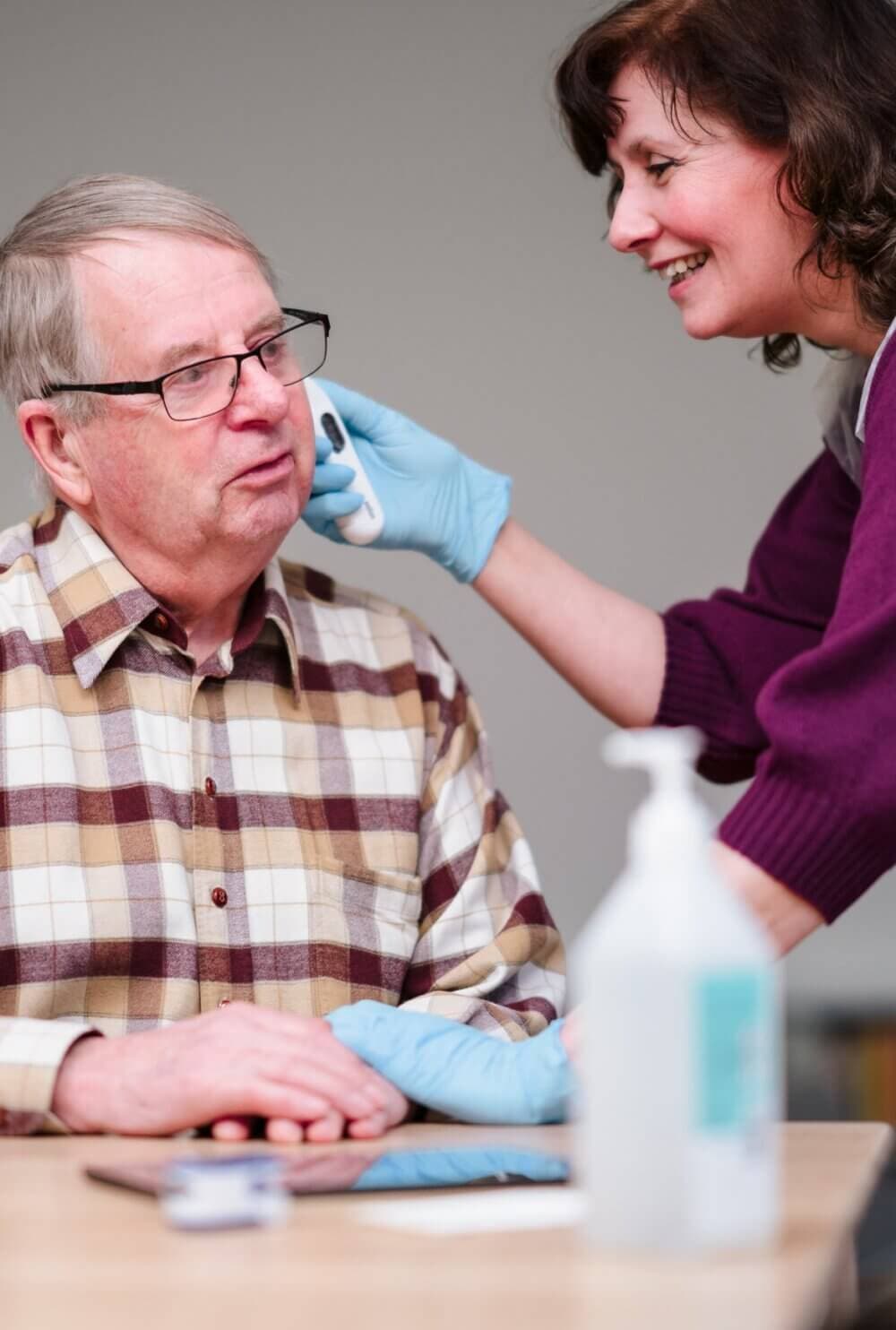 A caregiver in gloves wipes an elderly man's face with a cloth while smiling. A sanitizer bottle is on the table. - Home Instead