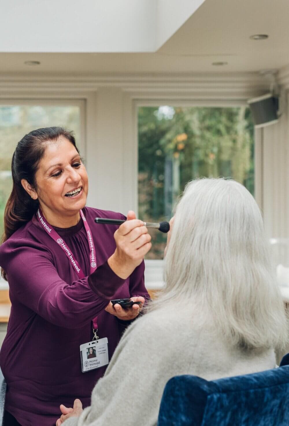 A Care Professional applying makeup to an elderly woman with white hair seated in a bright room with large windows. - Home Instead Poole