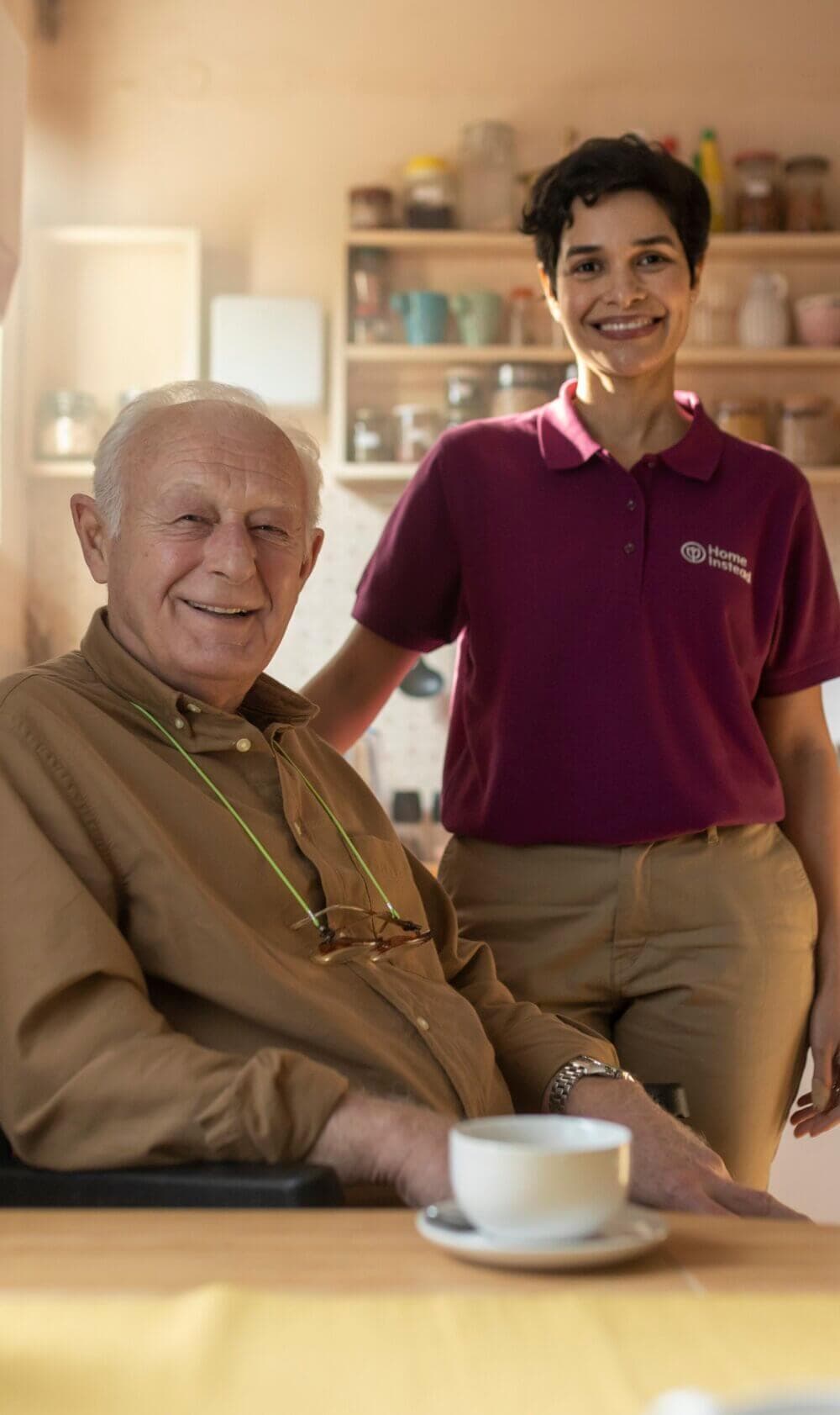 An elderly man sitting at a table with a cup of coffee, next to a smiling caregiver in a purple shirt. - Home Instead
