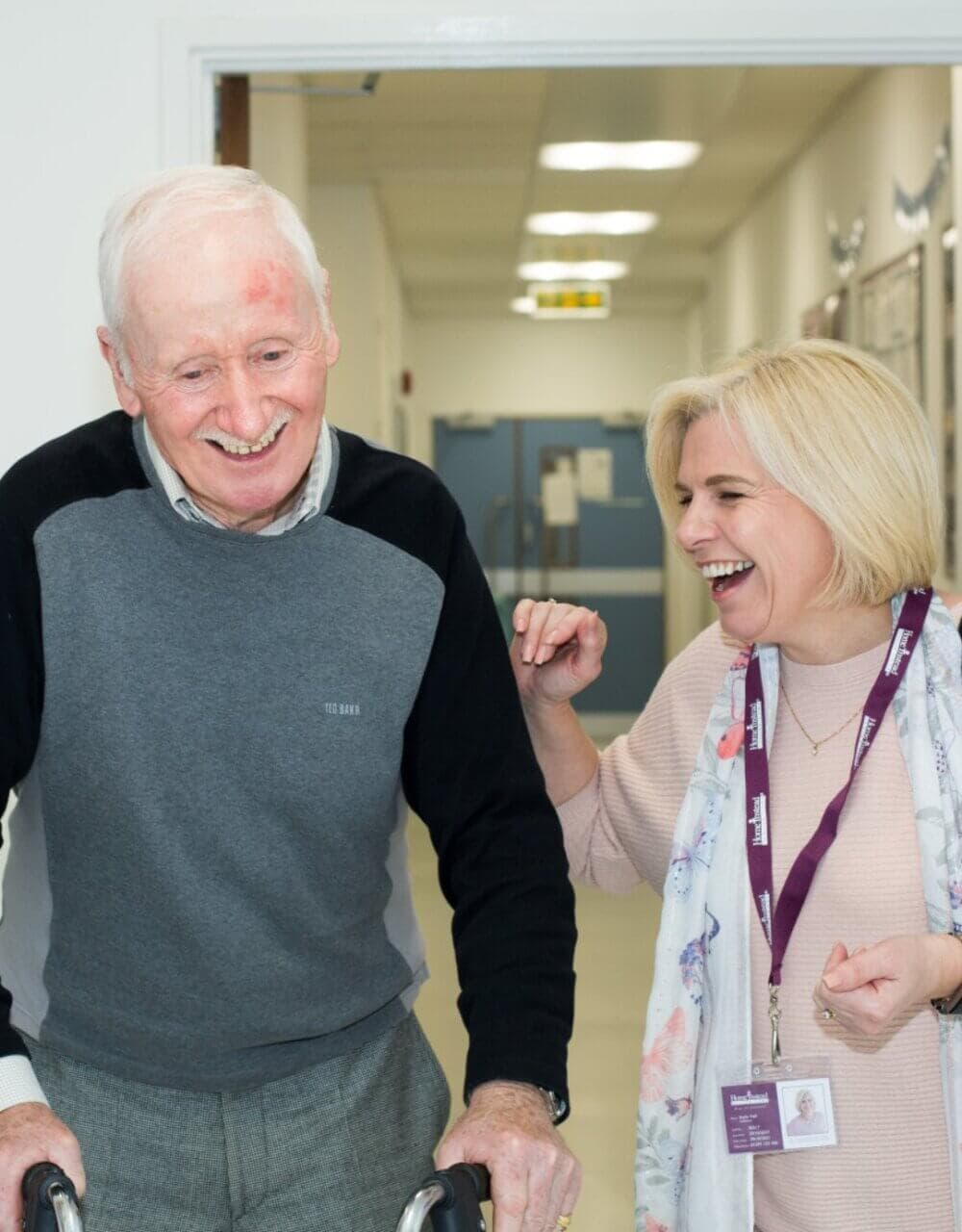 An elderly man using a walker and a woman laughing together in a hallway. - Home Instead
