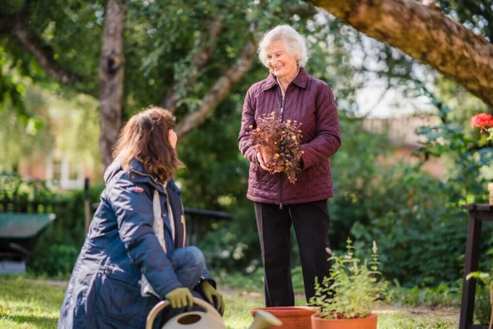 Elderly client holds plant while talking to younger woman kneeling in garden on a sunny day. Trees in the background. - Home Instead Bournemouth & Christchurch