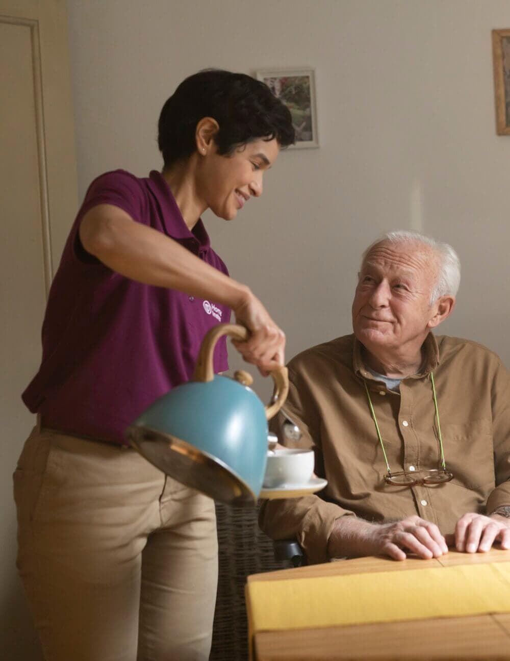 Elderly woman reading a book and smiling at a younger woman beside her in a kitchen setting. - Home Instead