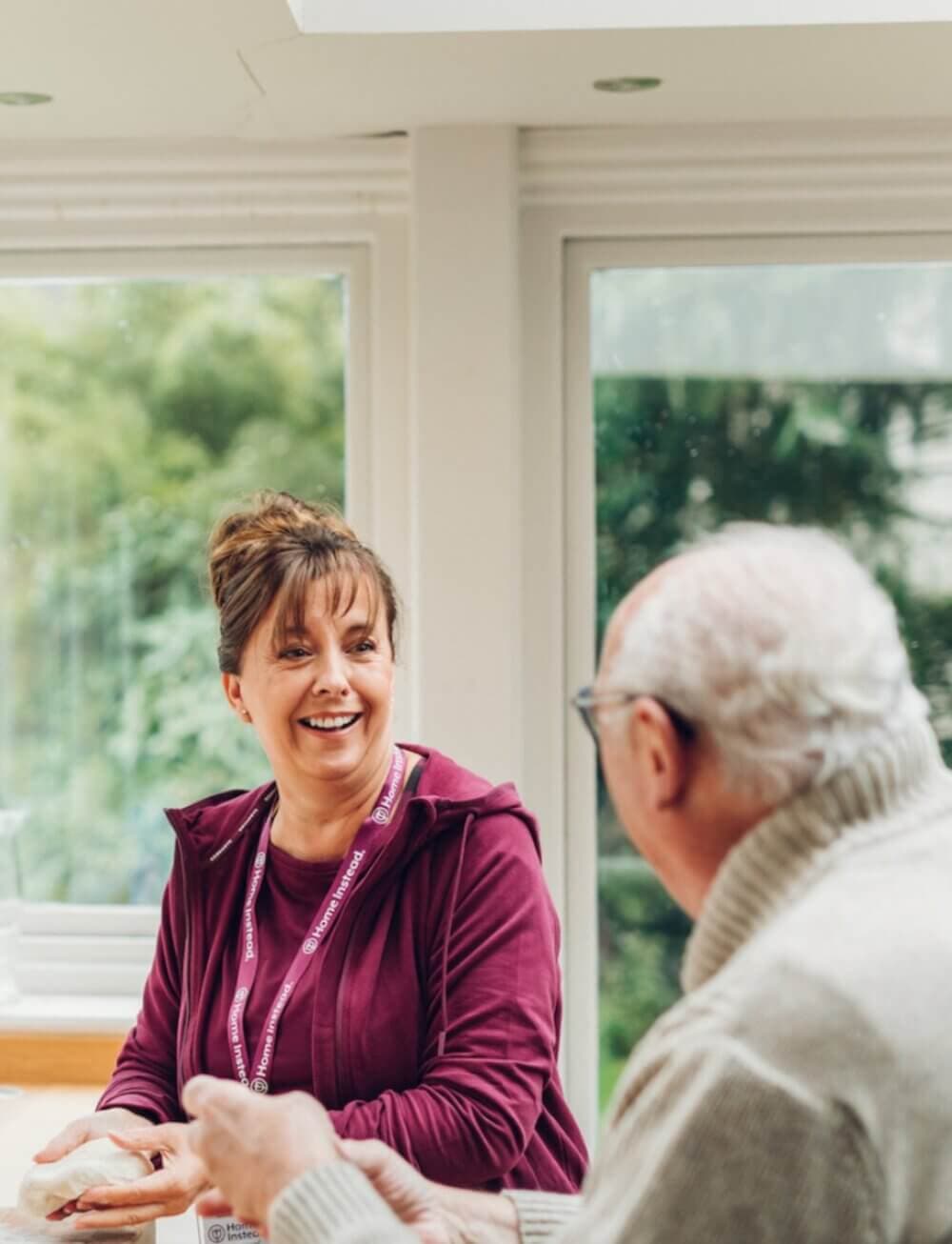 A woman and an elderly man smiling and talking at a table in a bright room with large windows. - Home Instead