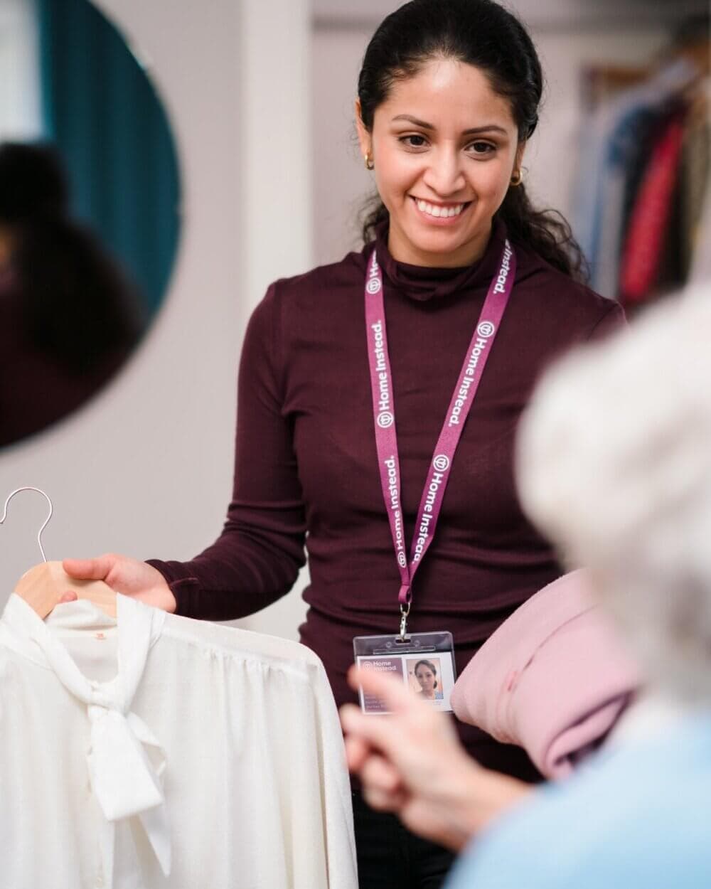 A woman with a staff badge assists a customer by holding up a white blouse and a pink sweater. - Home Instead