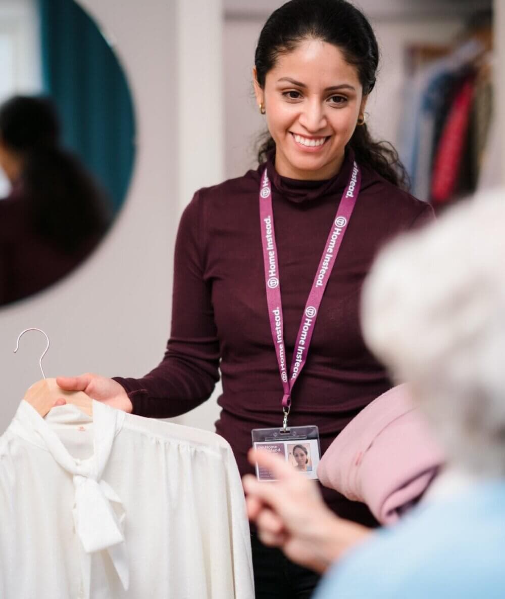 A woman with a name badge shows a white blouse on a hanger to another person in a room with clothes hanging in the background. - Home Instead
