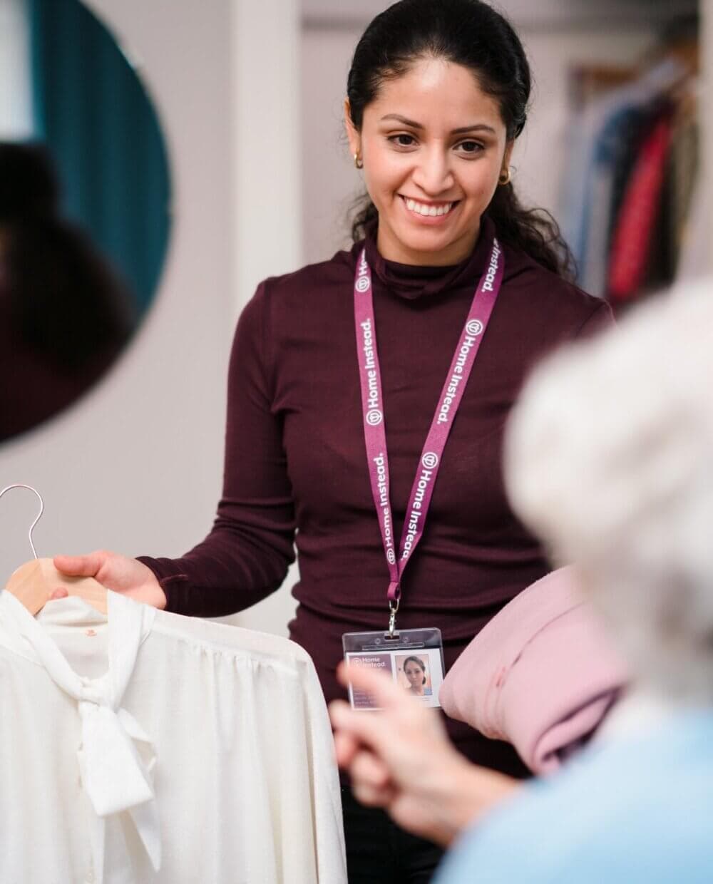 A woman helping another person choose clothes, holding a white blouse, wearing a name tag, and smiling warmly. - Home Instead