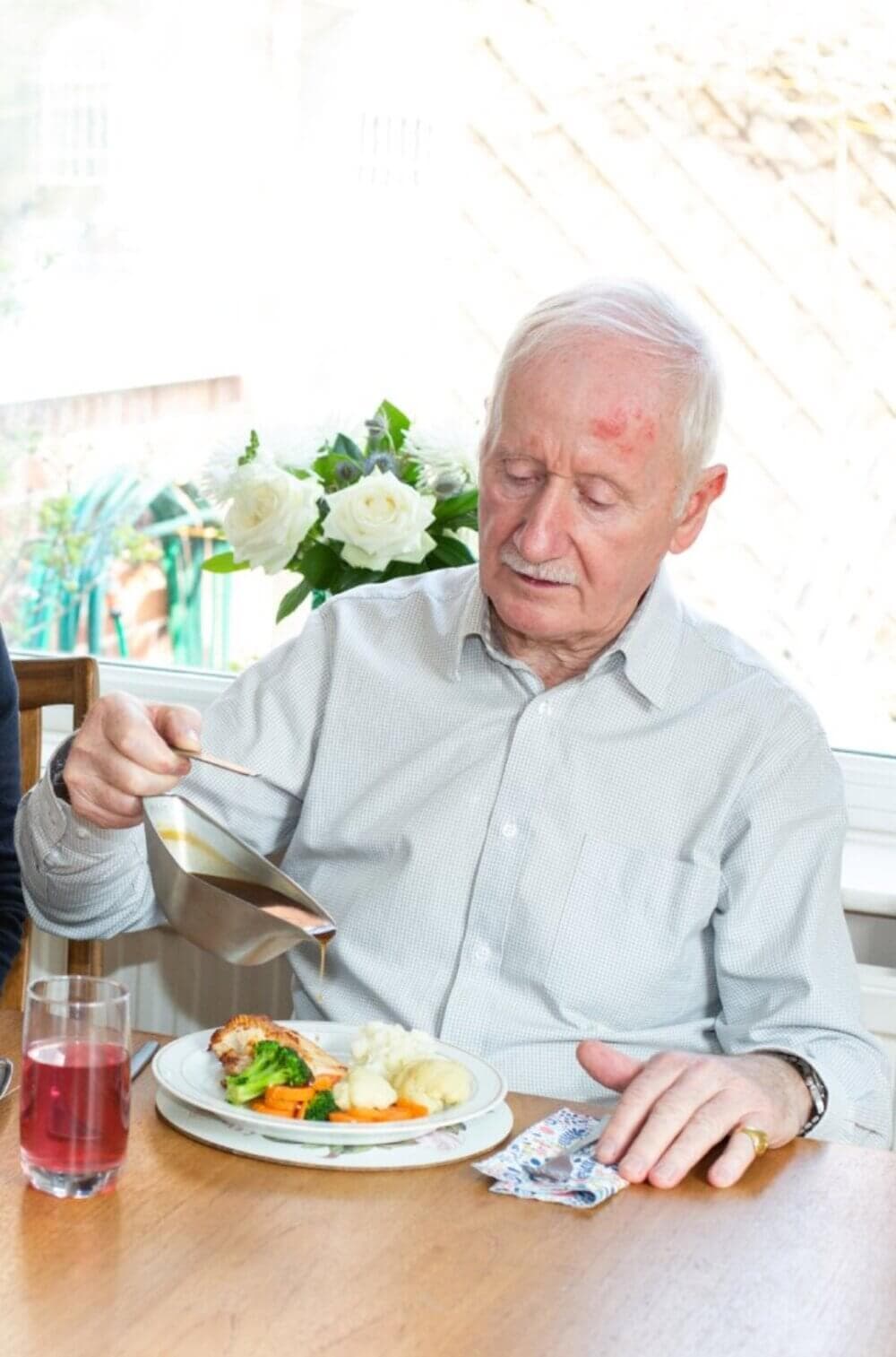 An elderly man in a gray shirt pours sauce over vegetables on his plate at a dining table. - Home Instead