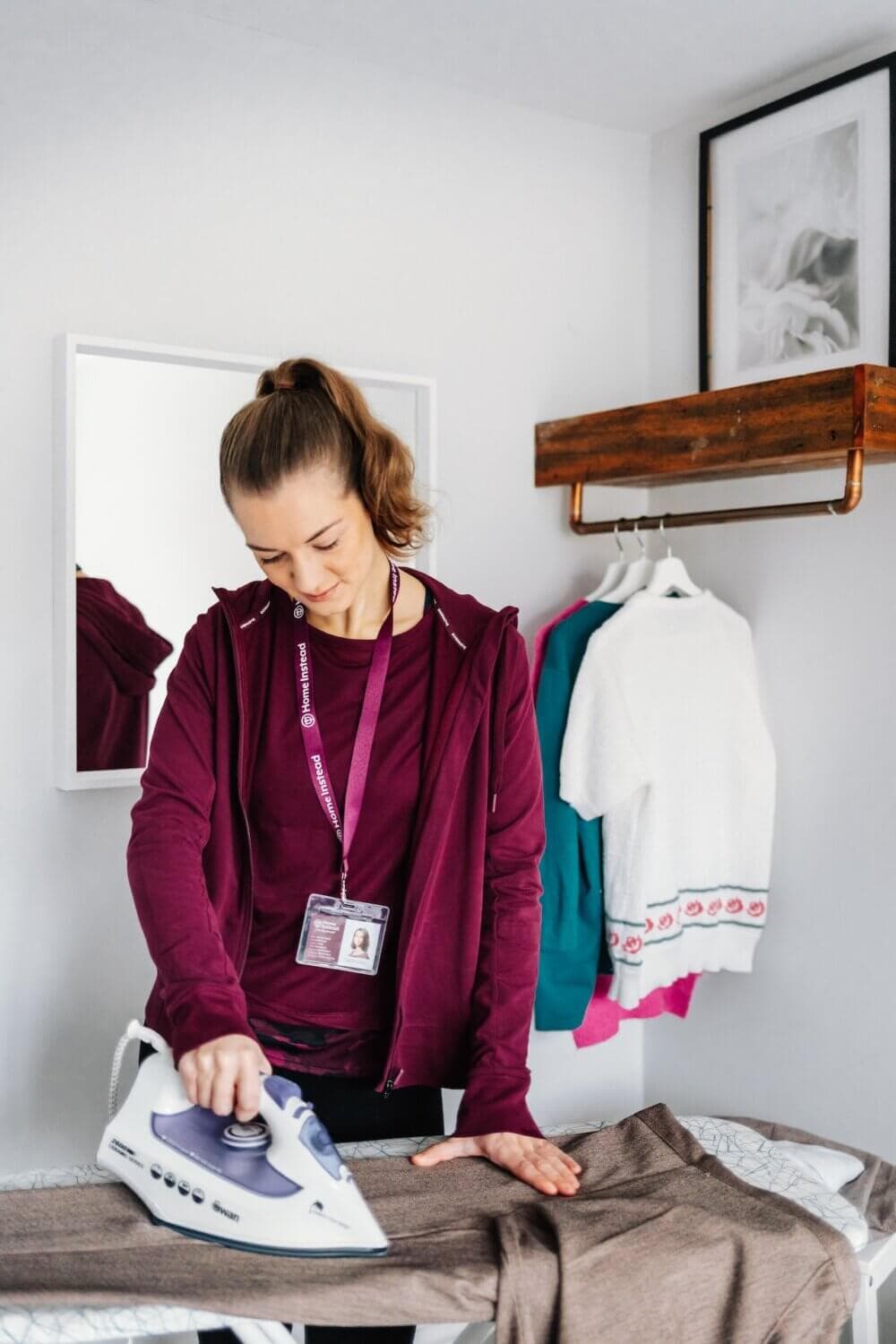 Person ironing a pair of pants in a room with a mirror, wall shelf, and clothing on hangers in the background. - Home Instead