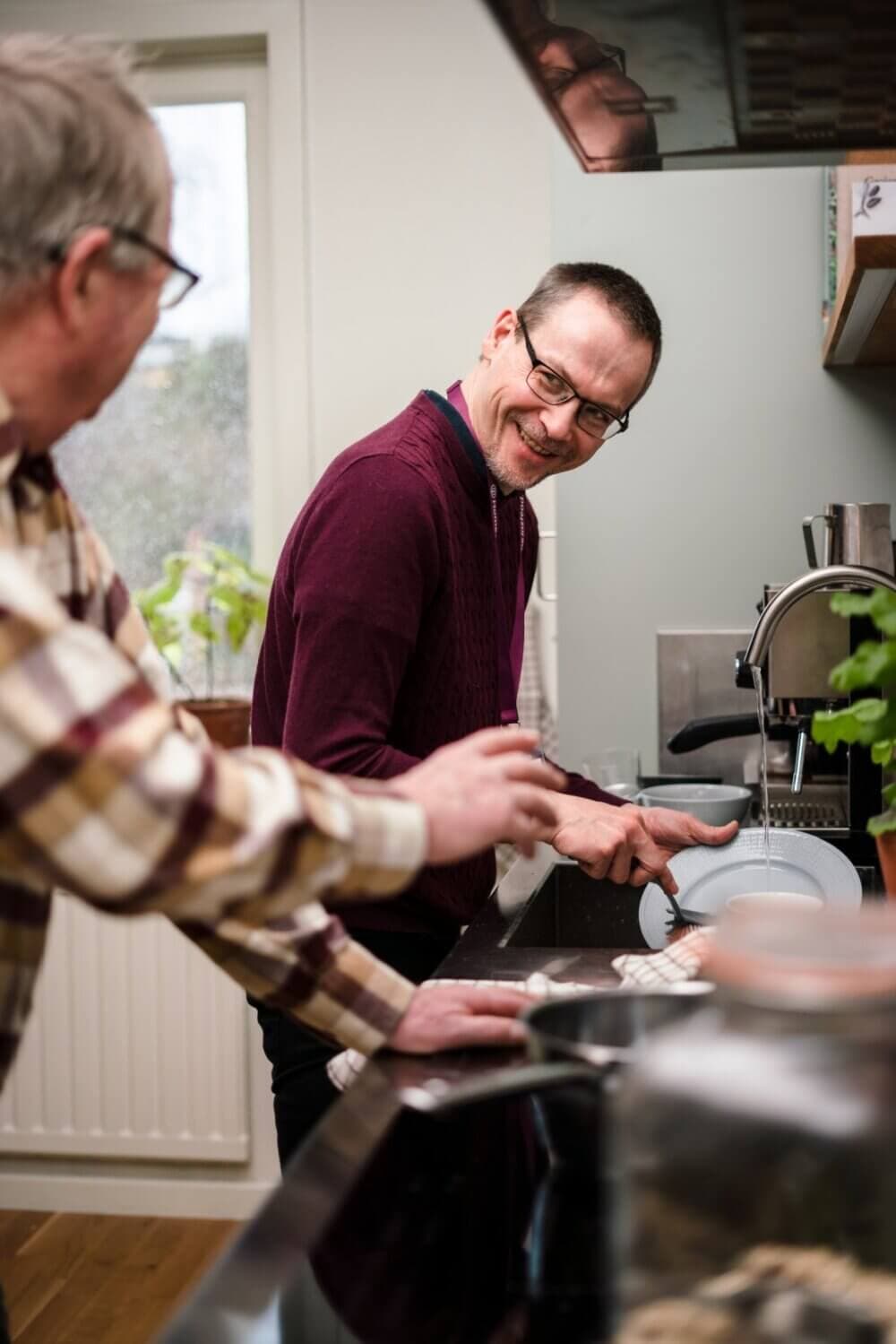 Two men, one washing dishes, the other speaking to him, in a kitchen with plants and a window in the background. - Home Instead