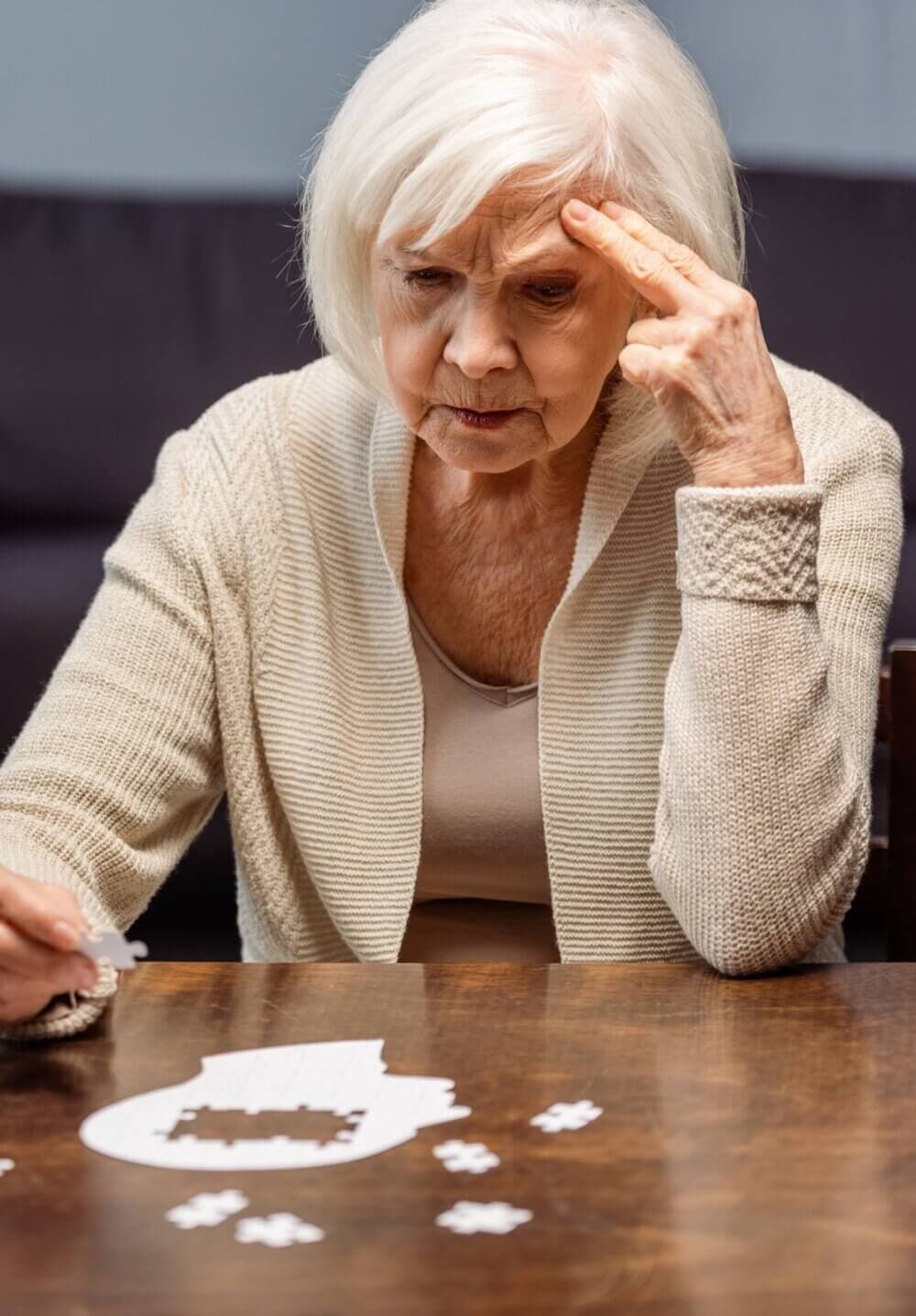 An elderly woman frowning, trying to fit a piece into a puzzle shaped like a human head at a wooden table. - Home Instead Southampton