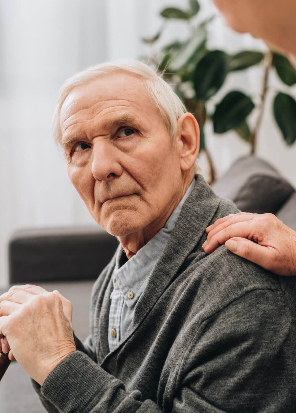 Elderly man sitting and looking up with a gentle hand on his shoulder. - Home Instead