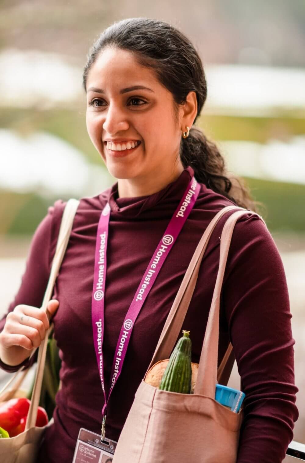 Smiling person carrying bags of groceries, wearing a maroon top and a lanyard around their neck. - Home Instead