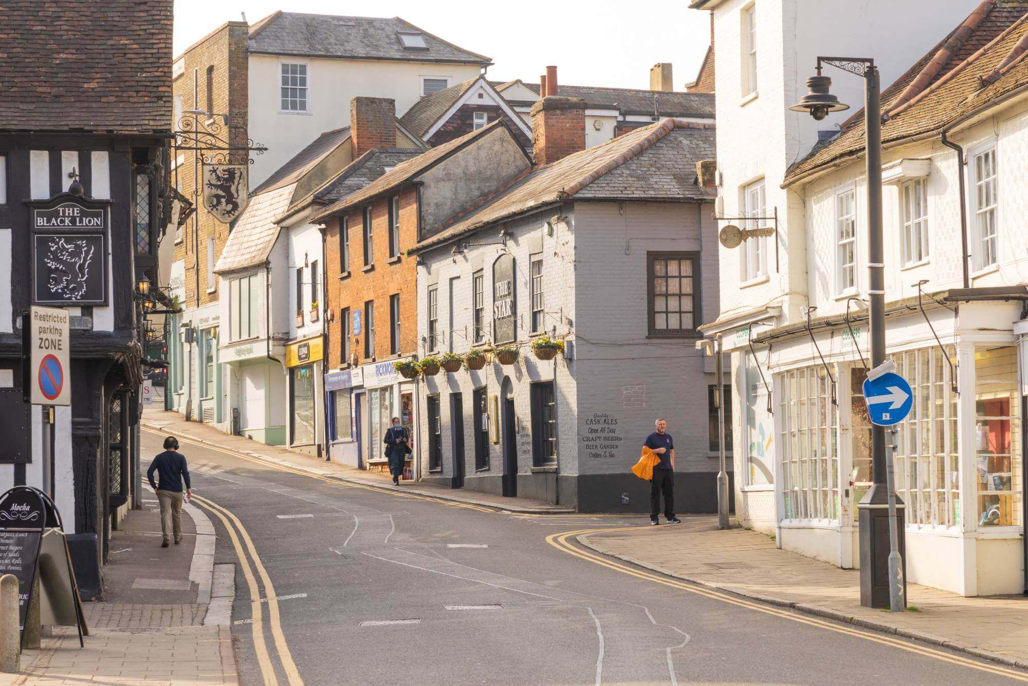 A narrow street in a small town with various buildings, a few pedestrians, and a street sign for The Black Lion pub. - Home Instead