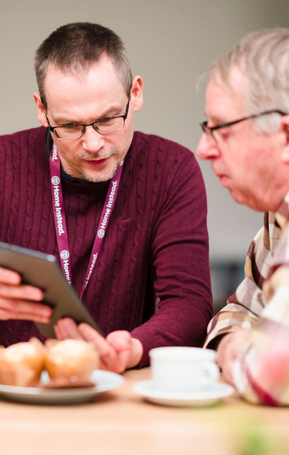 Two men, one showing the other a tablet, sit at a table with pastries and a cup of coffee. - Home Instead