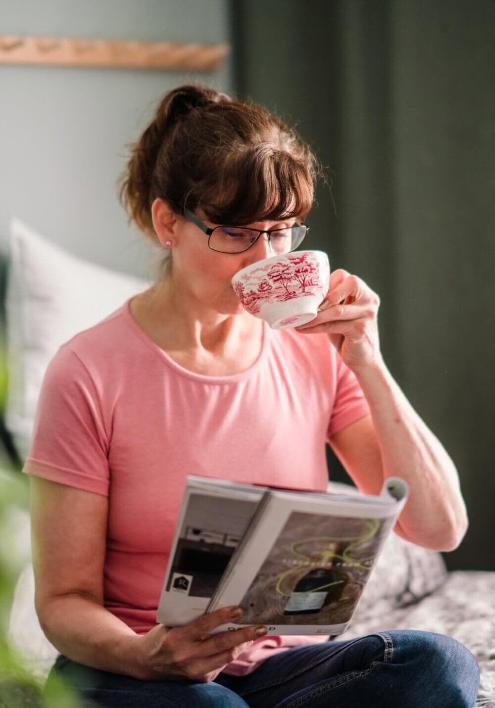 Woman in pink shirt reading a magazine and drinking from a floral-patterned teacup. - Home Instead