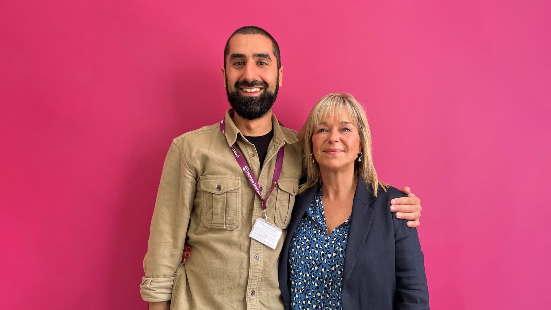 Two people standing together smiling in front of a bright pink background. The person on the left has an arm around the other. - Home Instead