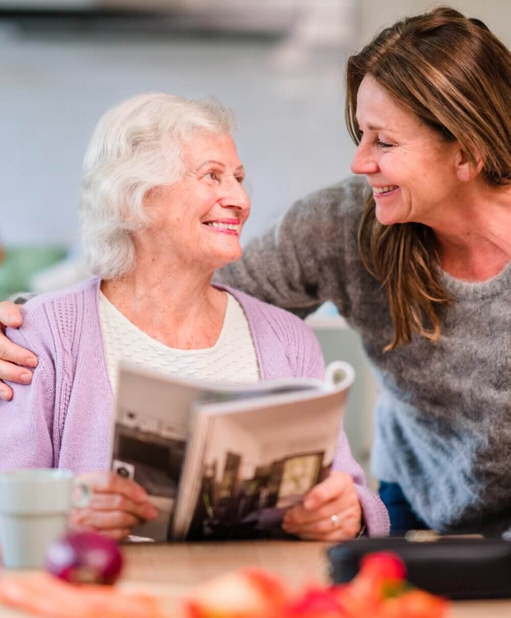Elderly woman holding a magazine, smiling at a woman beside her who has her arm around her. They are both smiling warmly. - Home Instead