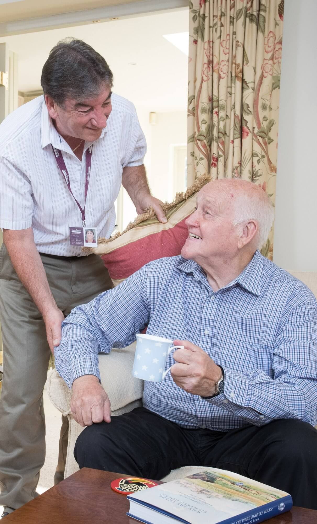 A caregiver assists an elderly man holding a mug, both smiling in a cozy living room setting. - Home Instead