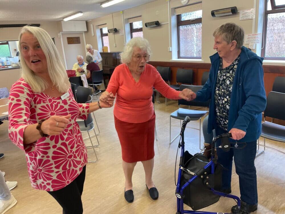 Three elderly women holding hands and dancing in a community room, with one using a walker. - Home Instead