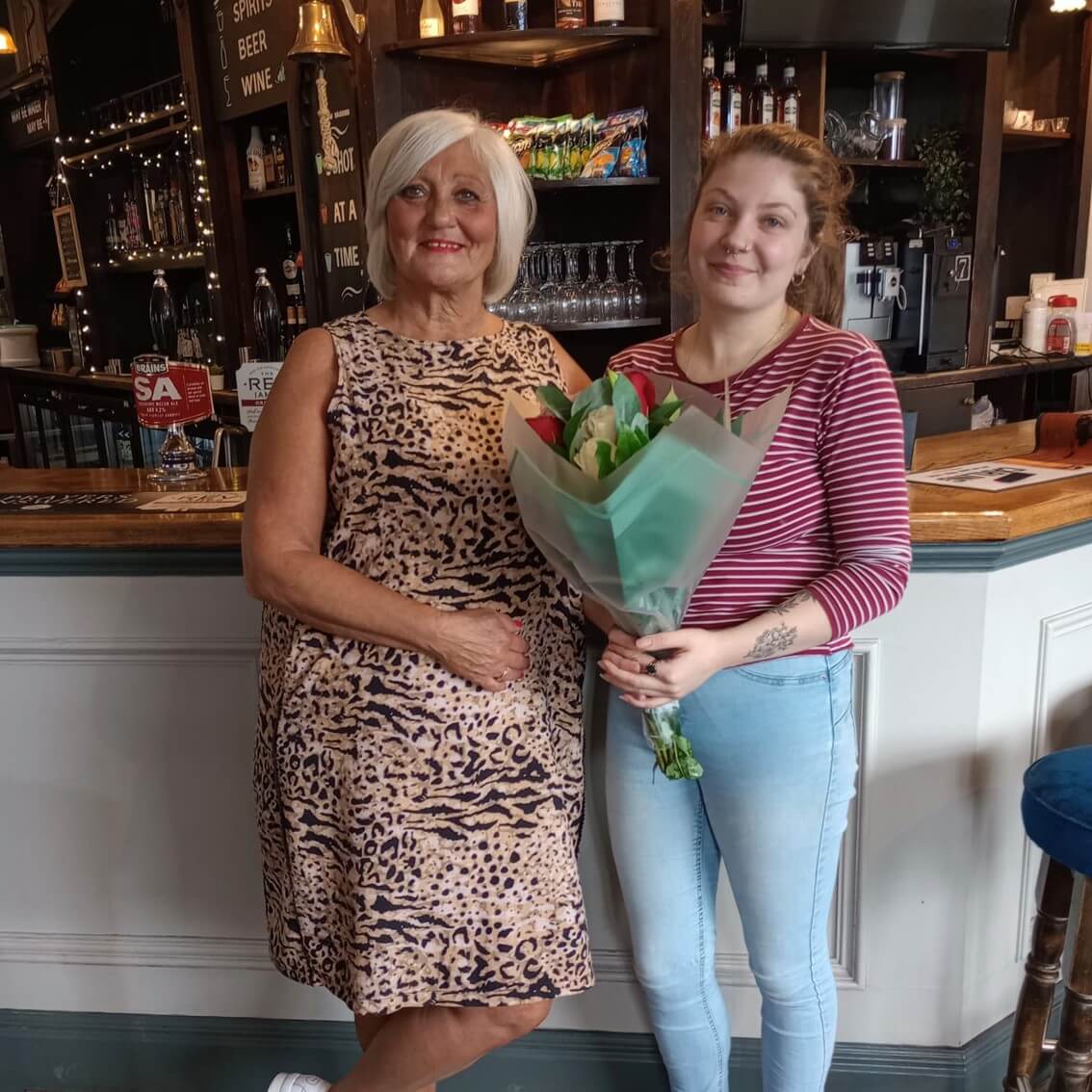Two women standing in a pub; one holds flowers. Background includes a bar with bottles and glasses on shelves. - Home Instead