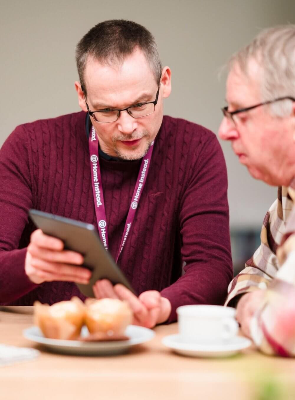 A man in a maroon sweater shows another man something on a tablet while seated at a table with muffins and a coffee cup. - Home Instead