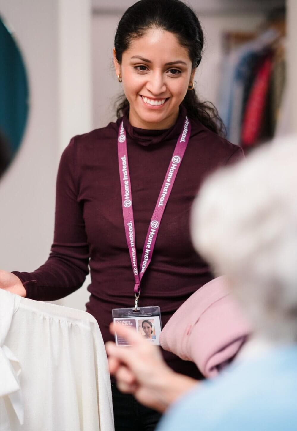 A smiling woman wearing a name badge and lanyard helps a customer with clothing in a store setting. - Home Instead