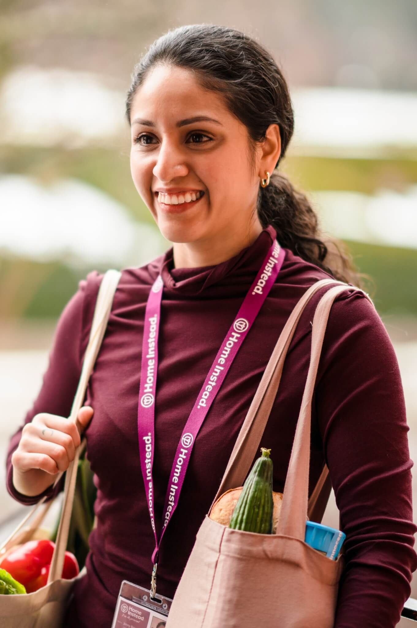 A smiling woman with a ponytail carries groceries in reusable bags, wearing a maroon shirt and a lanyard. - Home Instead