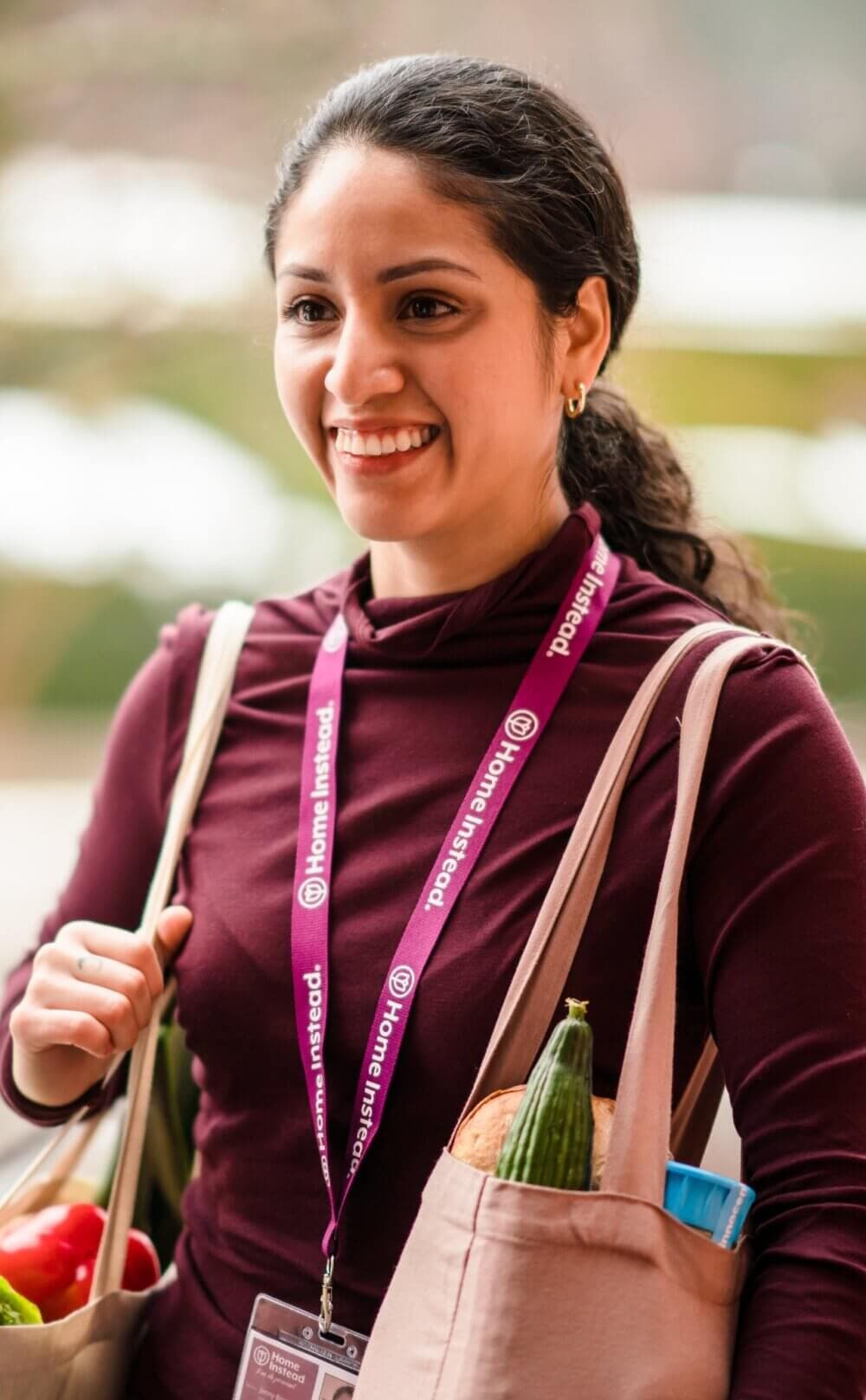 A smiling woman carrying bags of groceries, wearing a maroon shirt and a lanyard with the text "Home Instead". - Home Instead
