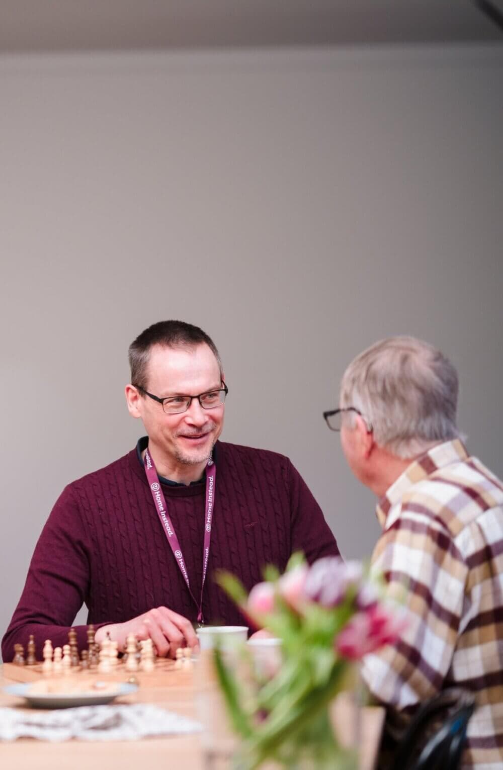 Two men sitting at a table, one wearing glasses and a lanyard, engaged in conversation. A bouquet of flowers in the foreground. - Home Instead