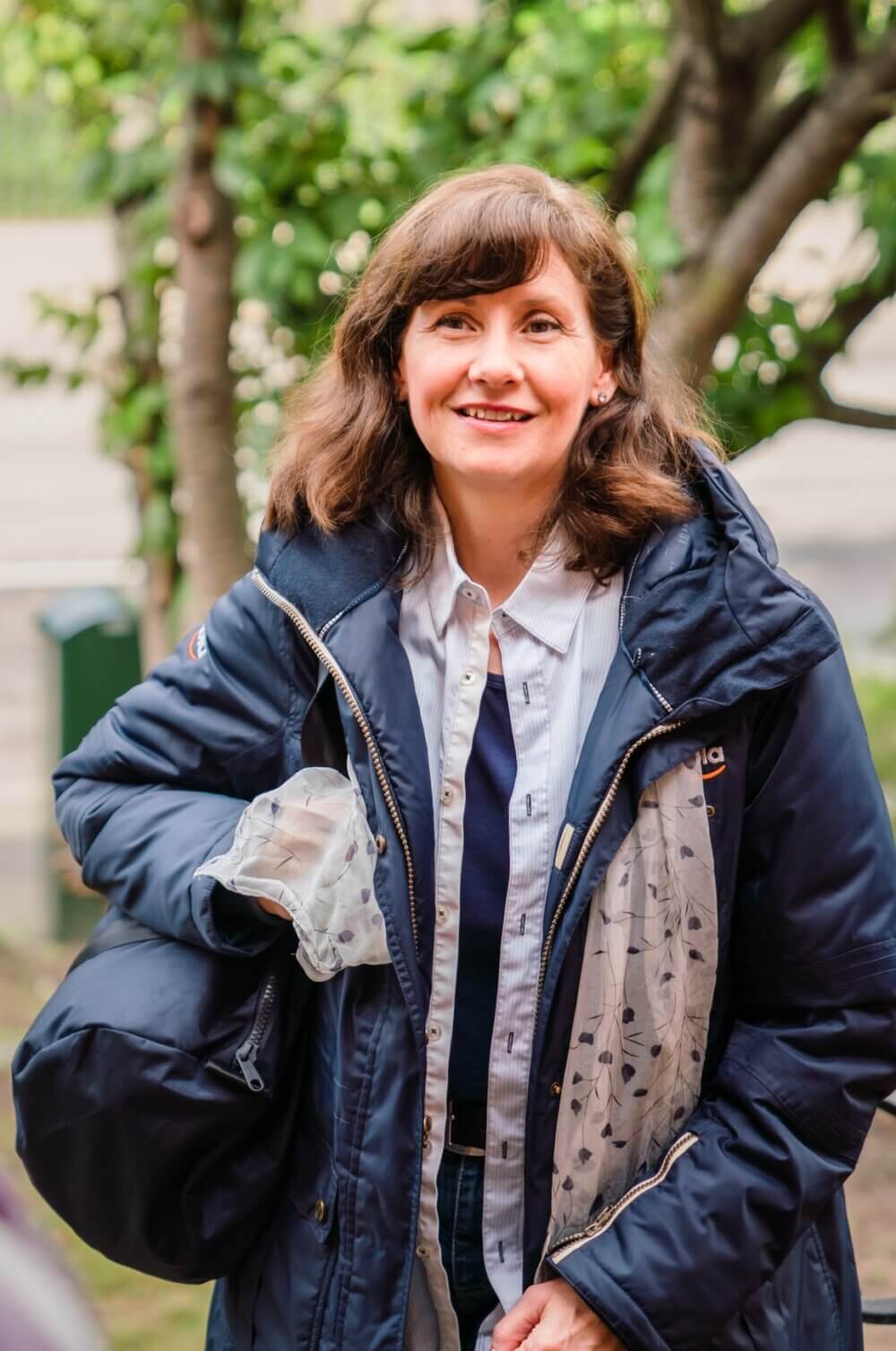 A smiling woman with brown hair wears a navy jacket and holds a plastic bag outdoors with trees in the background. - Home Instead