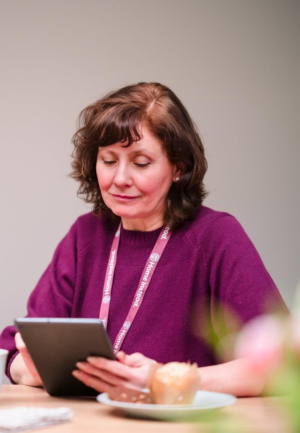 Woman in purple sweater reading on a tablet, wearing a lanyard, seated at a table with a plate and pastry. - Home Instead