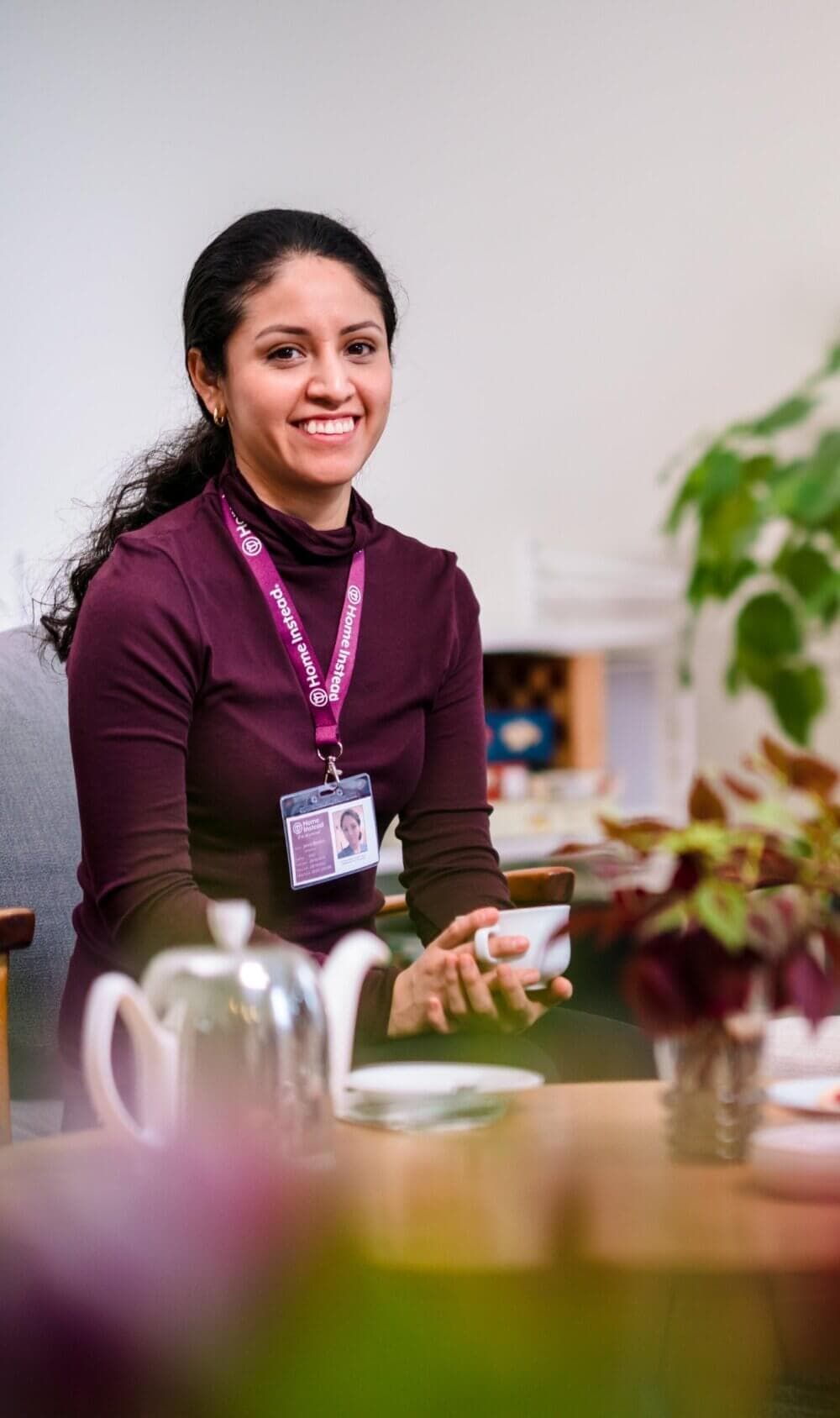 A smiling woman wearing a name badge sits at a table with a teapot, holding a teacup, and a plant in the foreground. - Home Instead