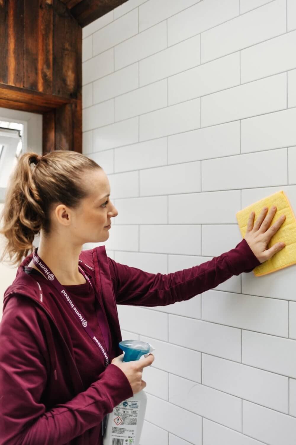 Person wearing a maroon hoodie cleaning white tiled wall with a yellow sponge and holding a spray bottle. - Home Instead Poole