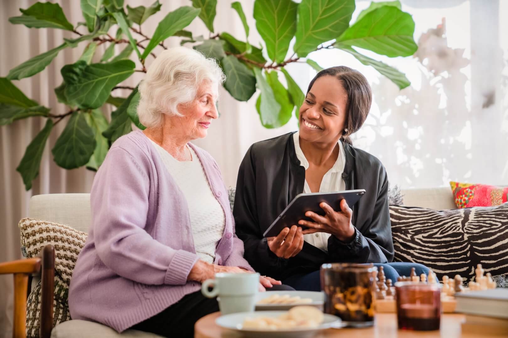 A woman holding a tablet and smiling while sitting on a couch with an elderly woman in a cozy living room. - Home Instead